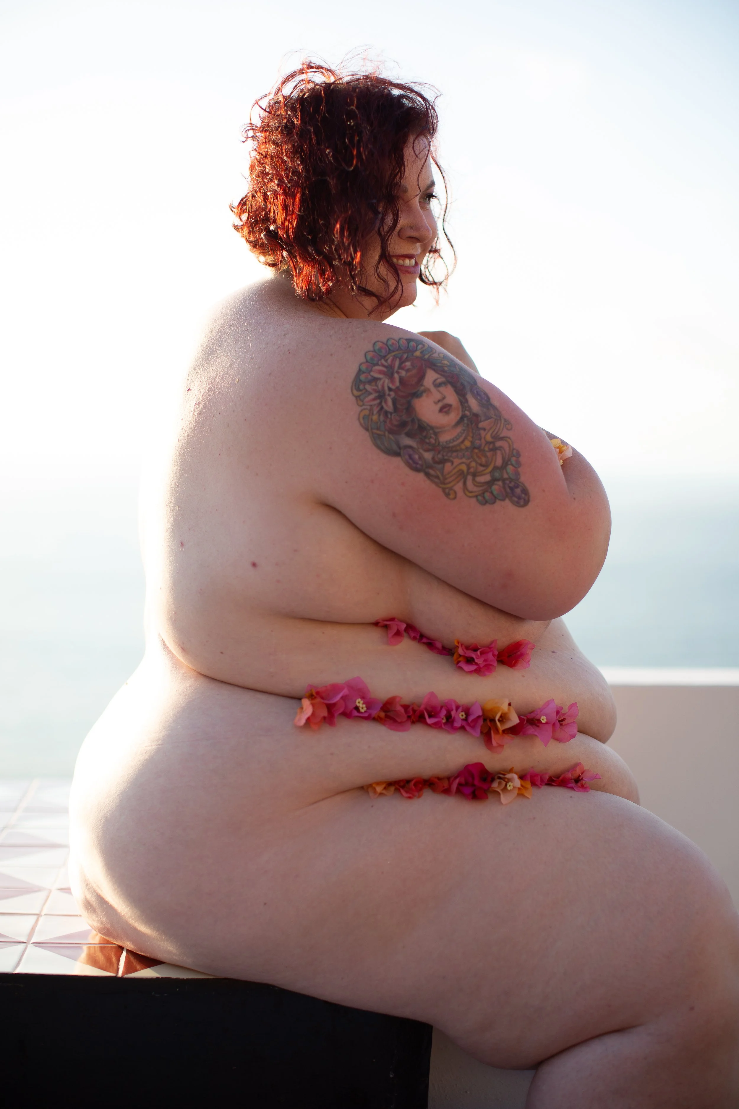 A woman with curly auburn hair, smiling, sitting outdoors with a background of blue sky and ocean, decorated with pink and orange flowers along her body, showing a tattoo of a woman's face with flowers on her shoulder.