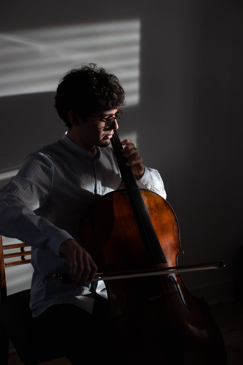 A man with curly hair and sunglasses playing a cello in a dimly lit room with sunlight streaming through window blinds.