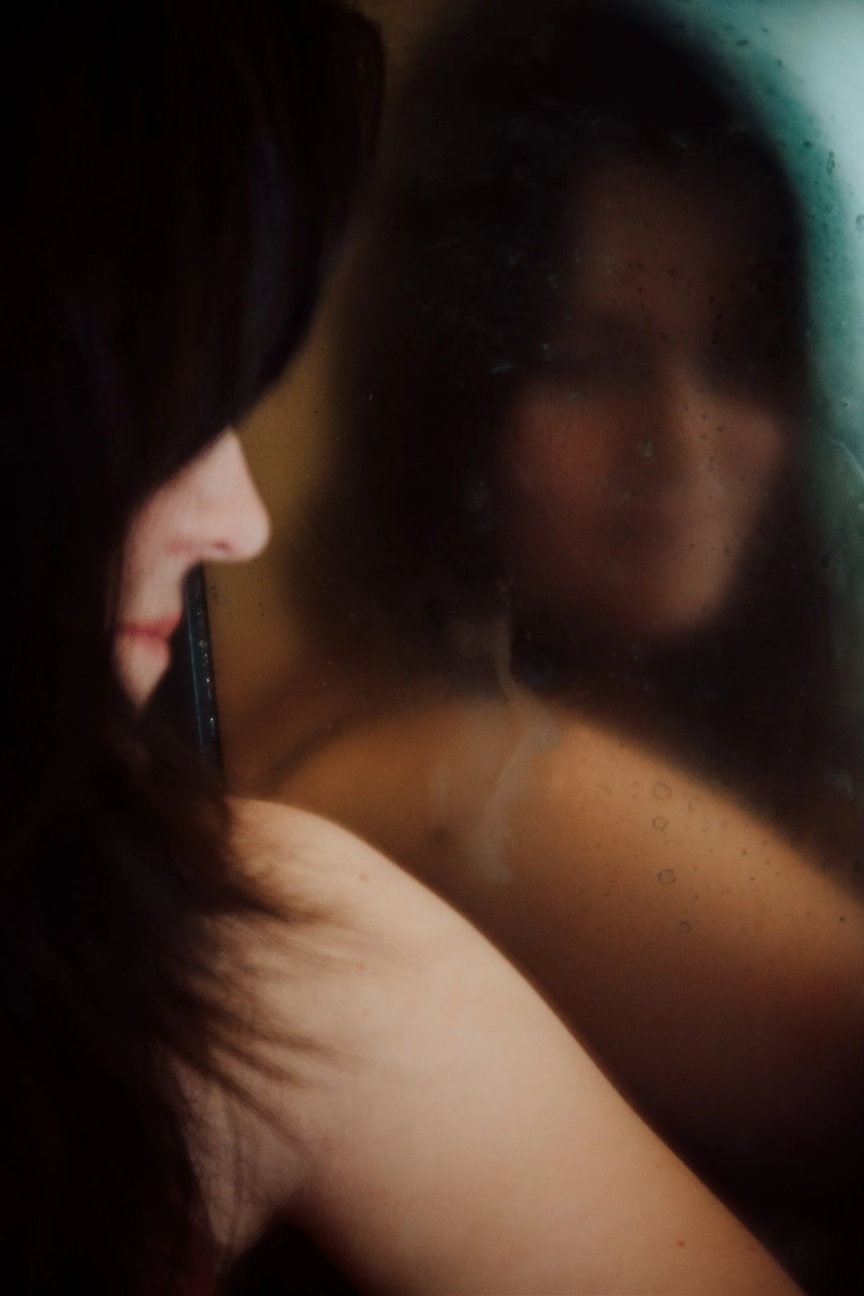 Close-up of a woman with dark hair and fair skin, smiling, with her face reflected in a mirror.