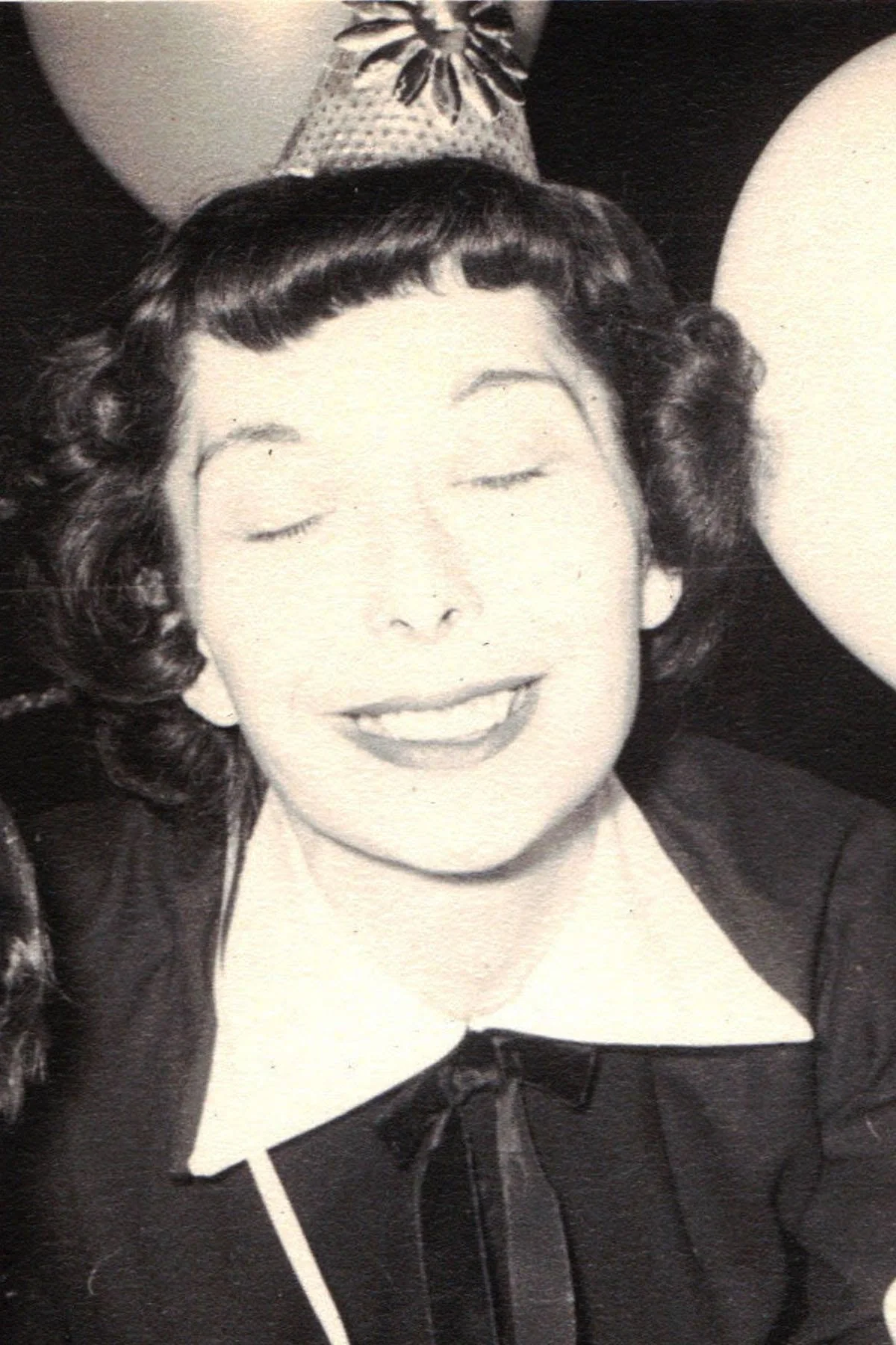 Black and white photo of a woman with short curly hair, wearing a smile, and a dark top with a bow detail at the neckline.