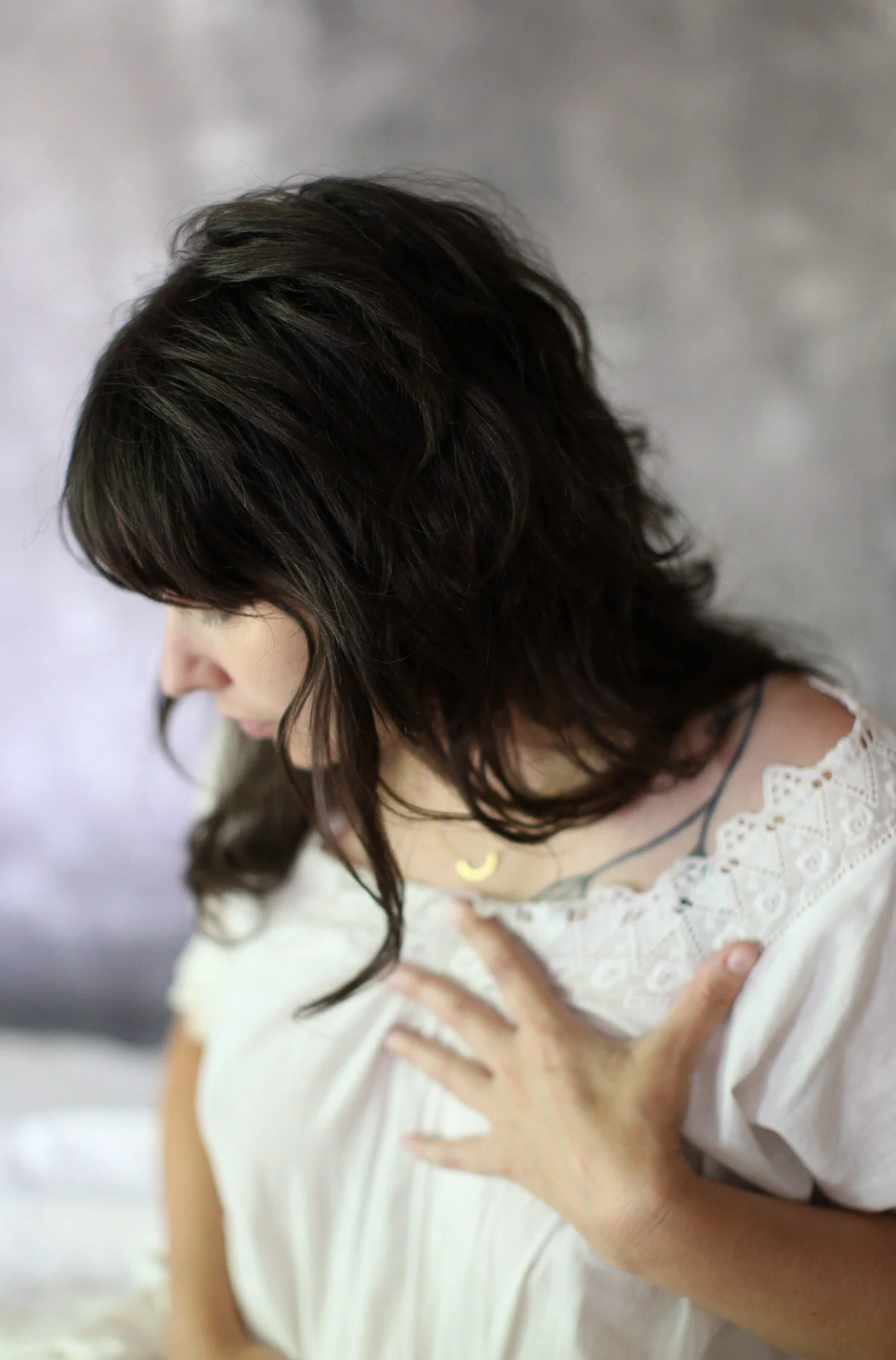 A woman with wavy, dark hair wearing a white dress with lace details, holding her chest with one hand, with a blurred, light-colored background.