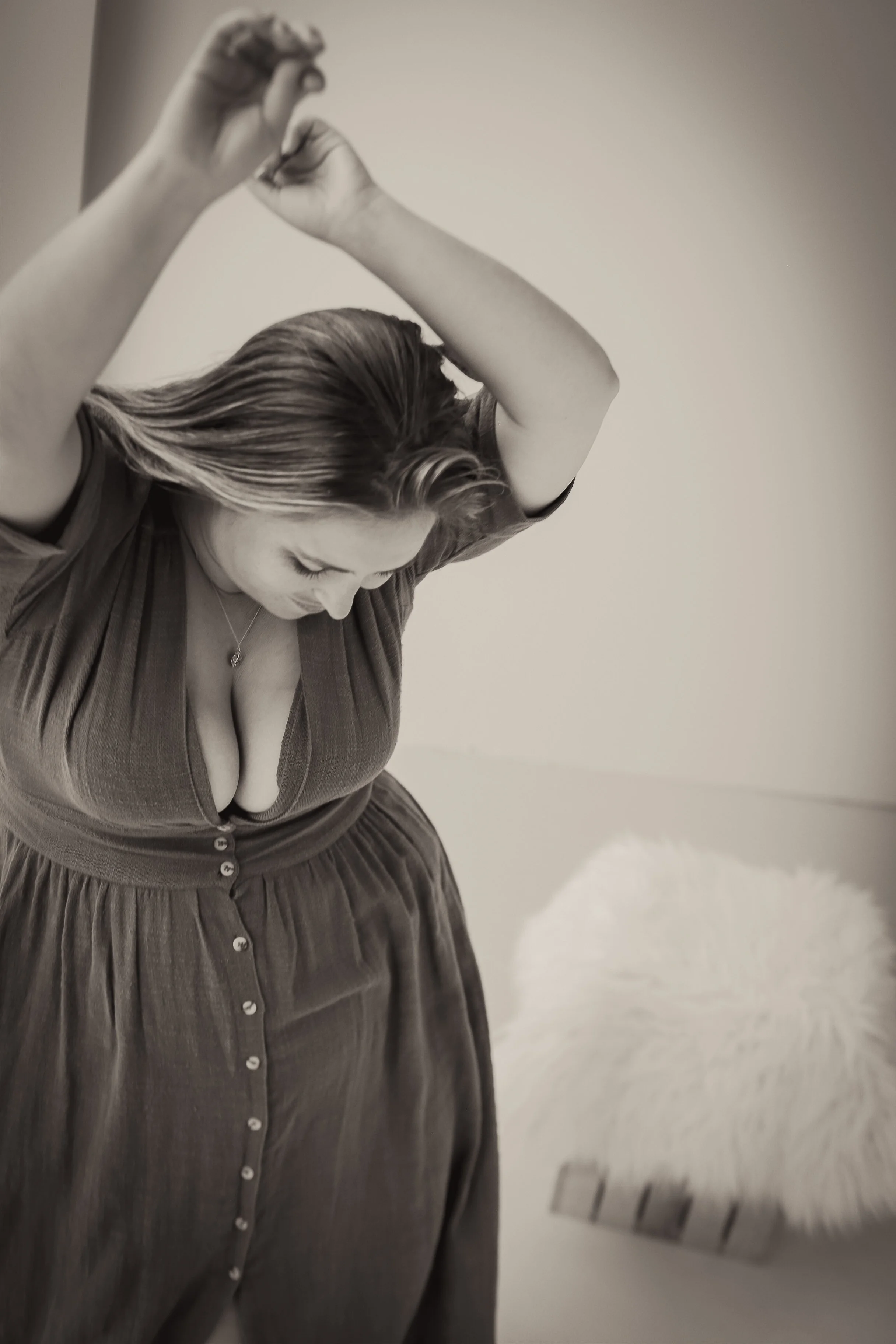 Black and white photo of a woman with long hair looking down, wearing a button-down dress with a deep neckline, in an indoor setting.