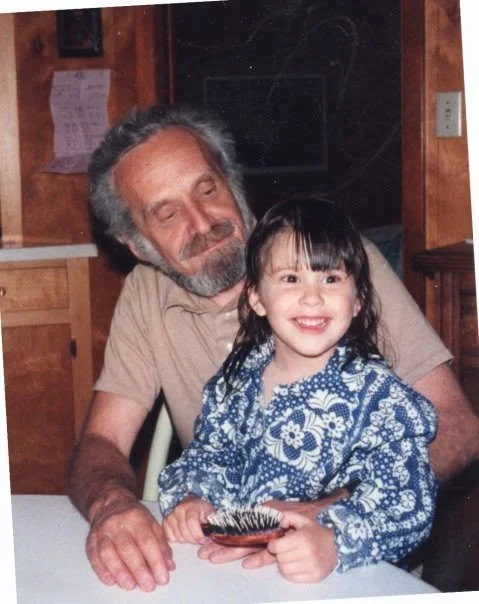 An elderly man with gray hair and a beard sitting at a table with a young girl with dark hair and bangs, smiling and holding a hairbrush, inside a wooden-paneled room.