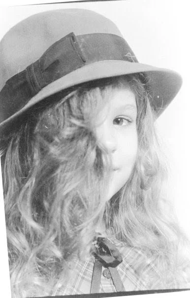 A young girl with curly hair wearing a wide-brimmed hat with a ribbon and a plaid shirt, smiling with one eye showing.