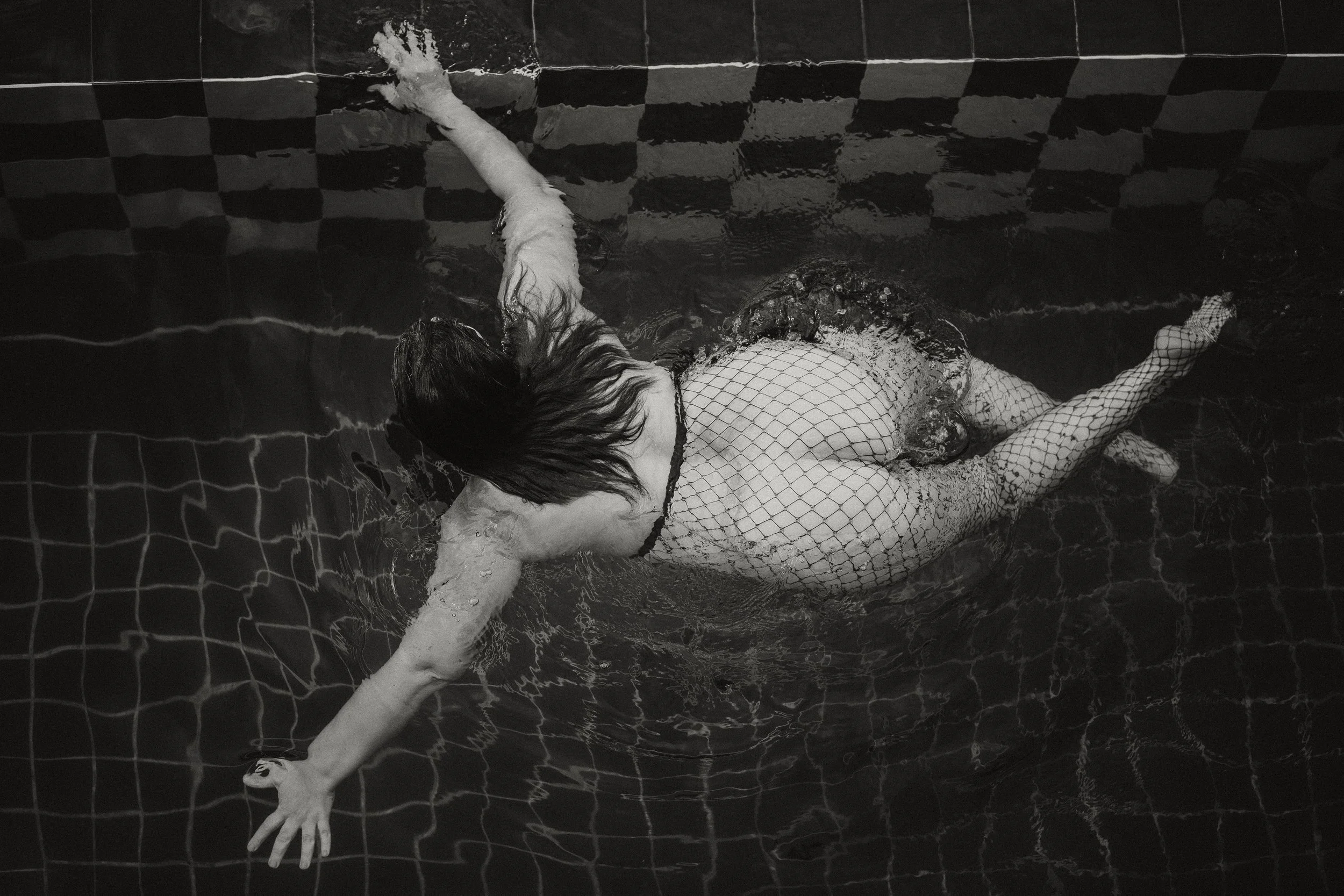 A person lying on their side in a swimming pool, wearing fishnet stockings and a patterned swimsuit, with long wet hair covering part of their face, near the edge of the pool with checkered tile tiles visible.