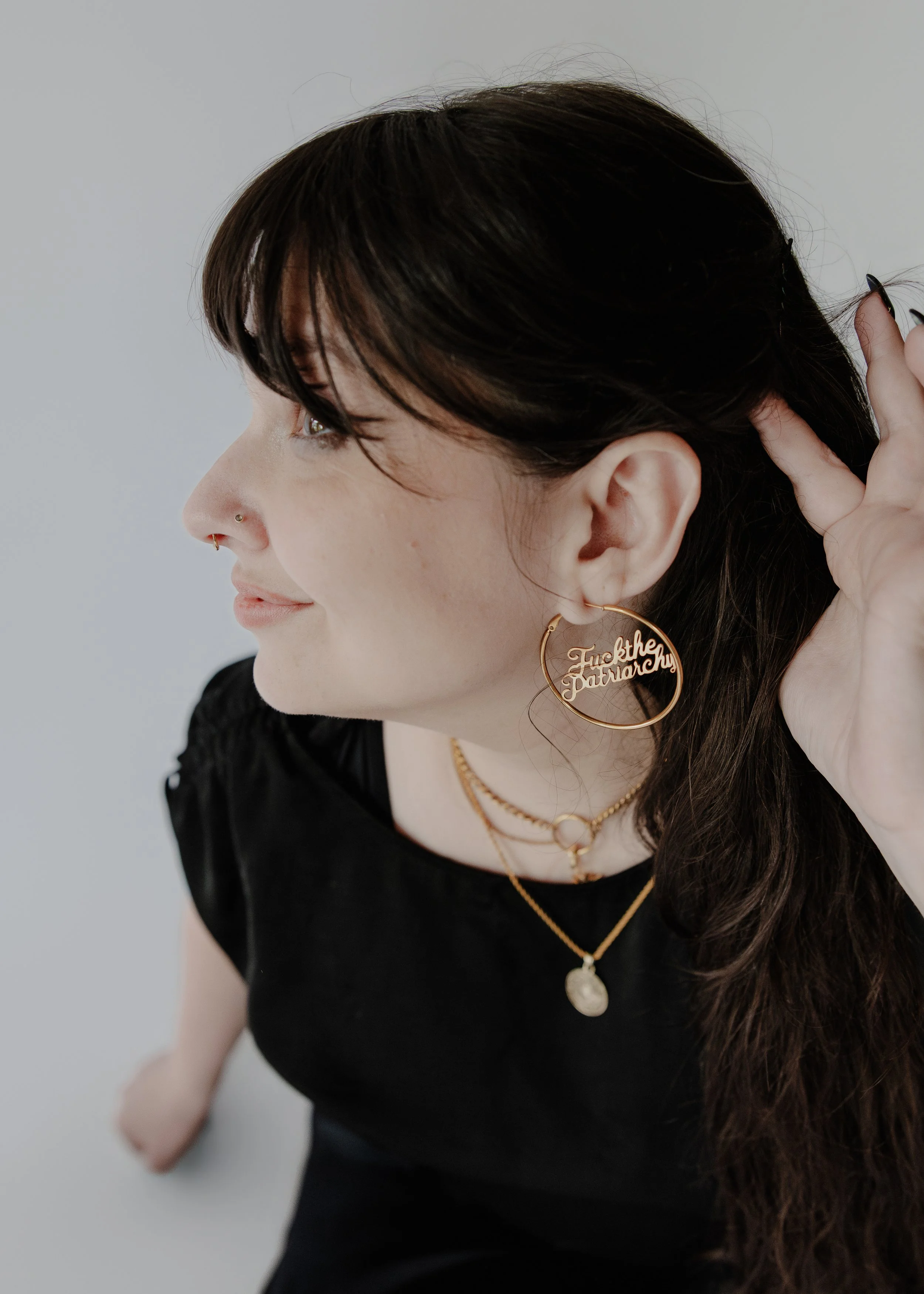 A woman with dark brown hair, gold earrings with a message, and layered necklaces, touching her hair with her right hand, wearing a black top, against a plain light background.