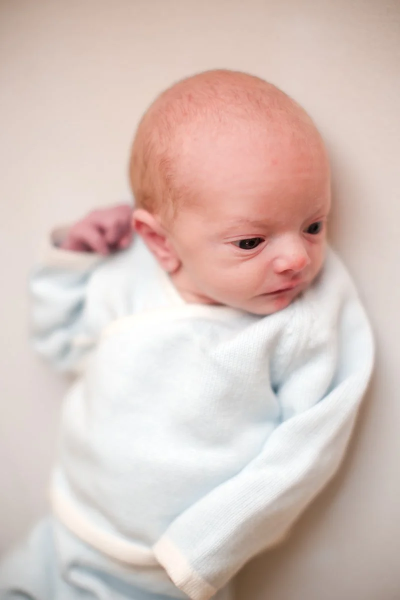 A newborn baby with a bald head, wearing a white outfit, lying on a light-colored surface, gazing to the side.