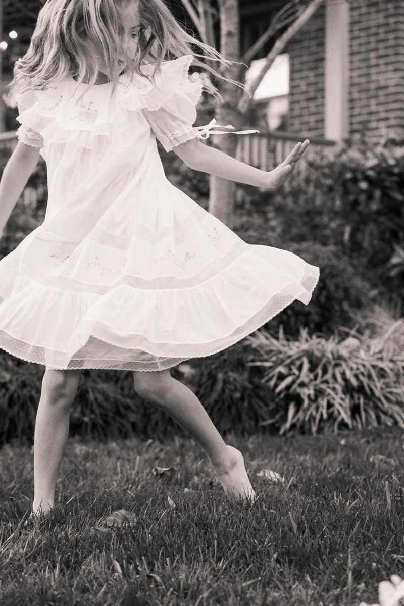 A young girl wearing a white dress spinning and twirling outdoors on a grassy lawn, with a house and plants in the background.