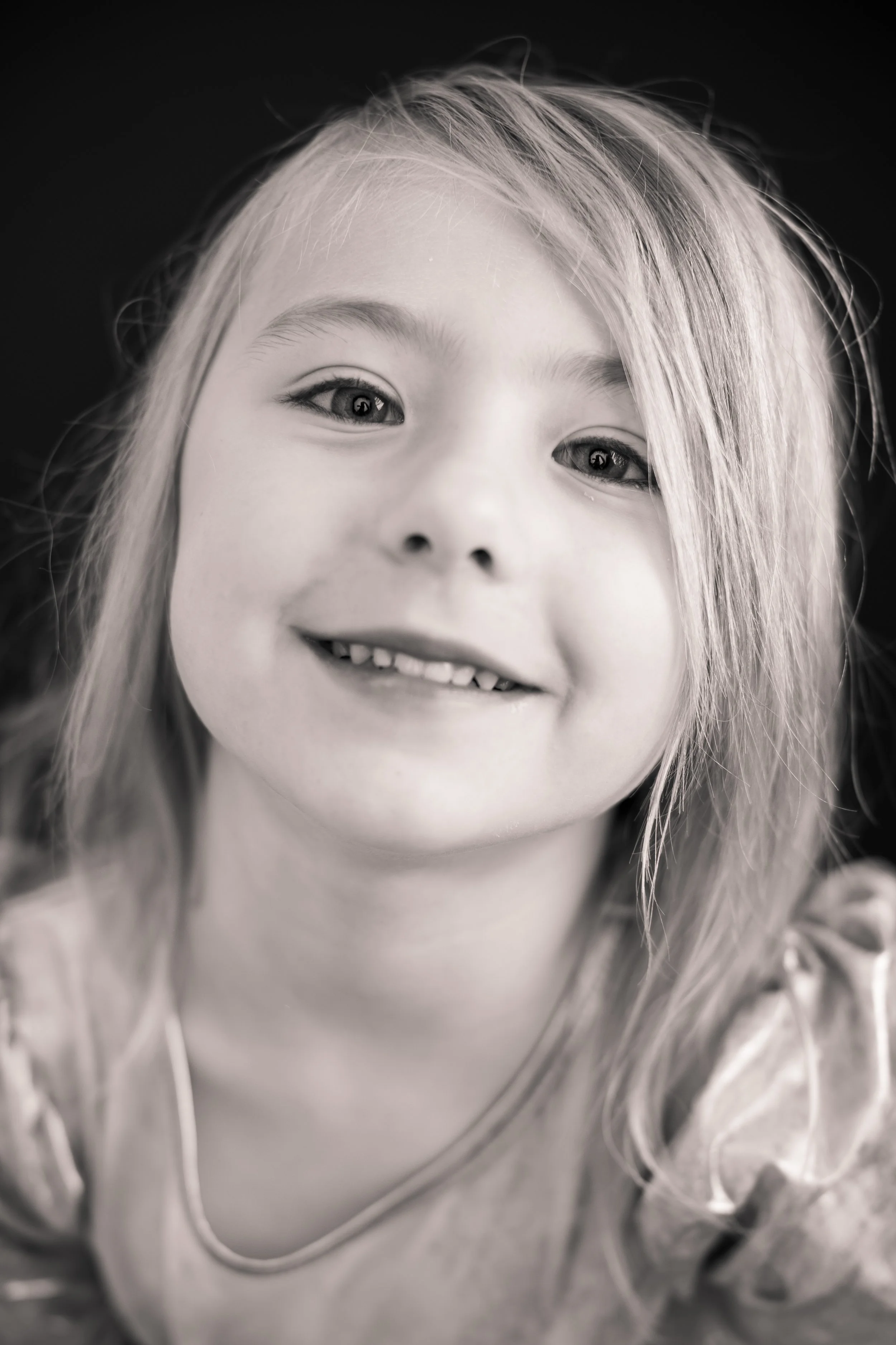Black and white close-up photo of a smiling young girl with light-colored hair, gleaming eyes, and a necklace.