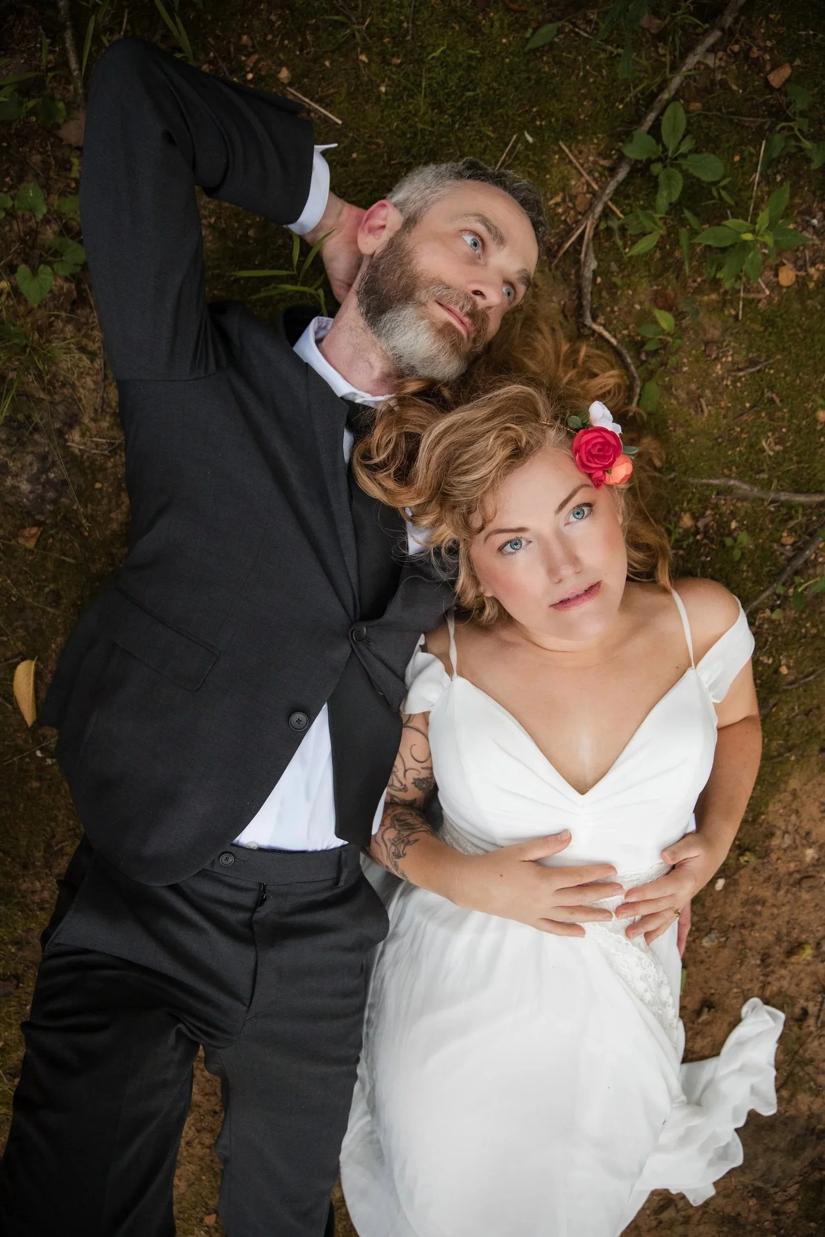 A man in a black suit and a woman in a white wedding dress lying on the ground outdoors among green plants and moss, looking up with relaxed expressions.