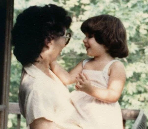 A woman and a young girl share a joyful moment indoors, with the woman holding the girl as they look at each other.
