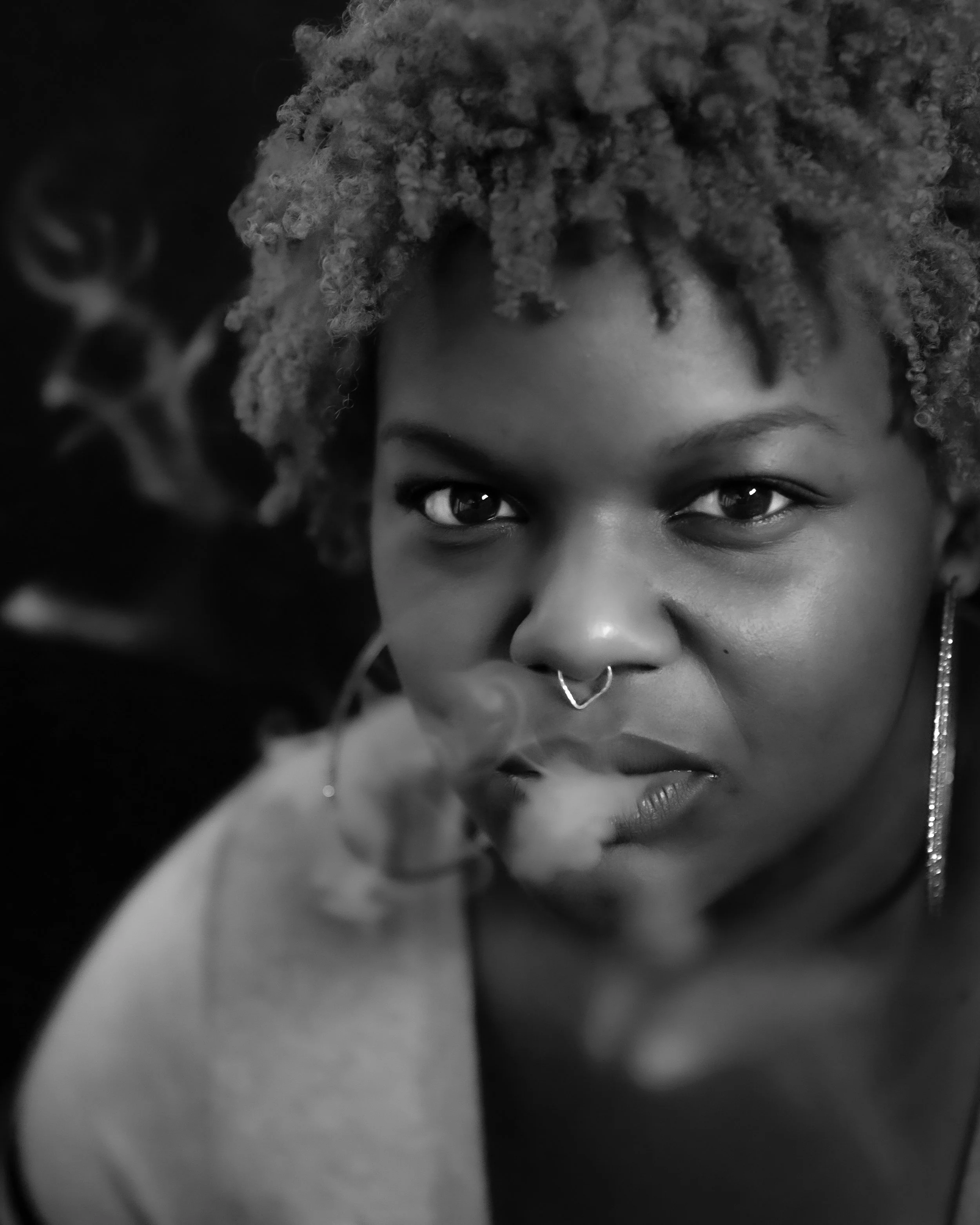 Close-up black-and-white photo of a person with curly hair, showing a portion of their face including eyes, nose, and lips, with a septum piercing.
