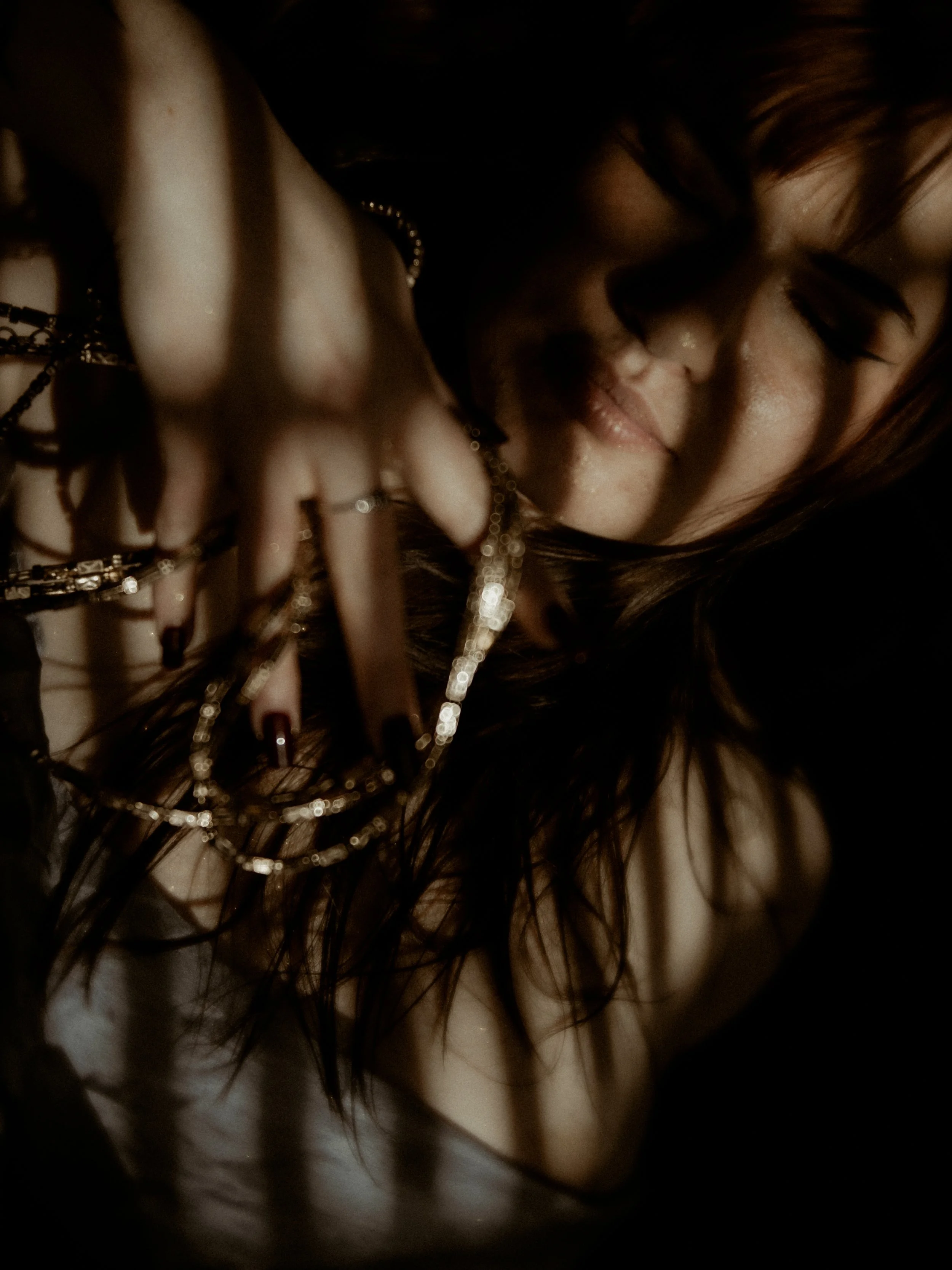 A close-up of a woman's face with wet hair and closed eyes, holding a chain with jewelry in front of her face in a dark setting.