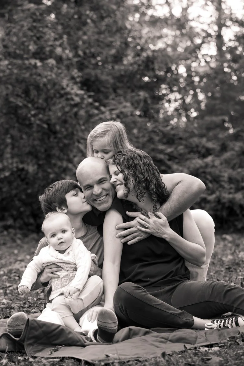 A black and white photo of a happy family outdoors, with a bald man smiling as he is hugged by a woman with curly hair, and surrounded by three children, including a baby, in front of a wooded background.