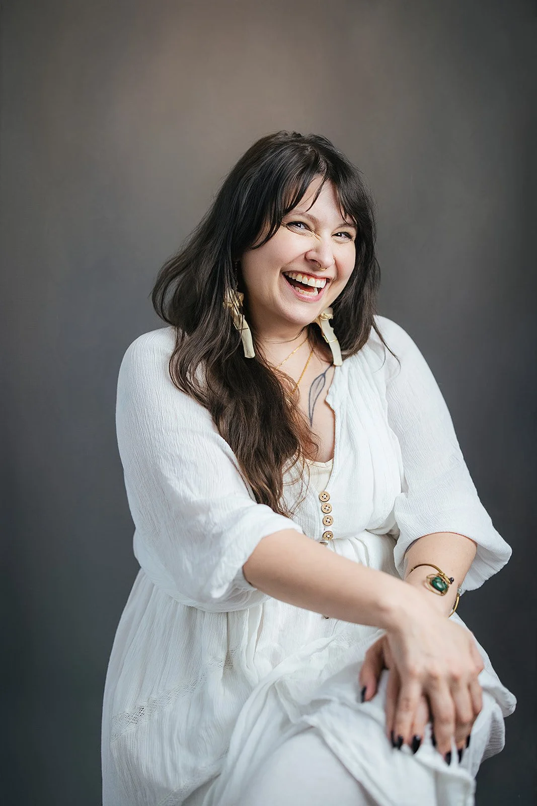 A woman with long dark wavy hair, wearing a white dress, smiling and showing her teeth, seated against a neutral gray background.