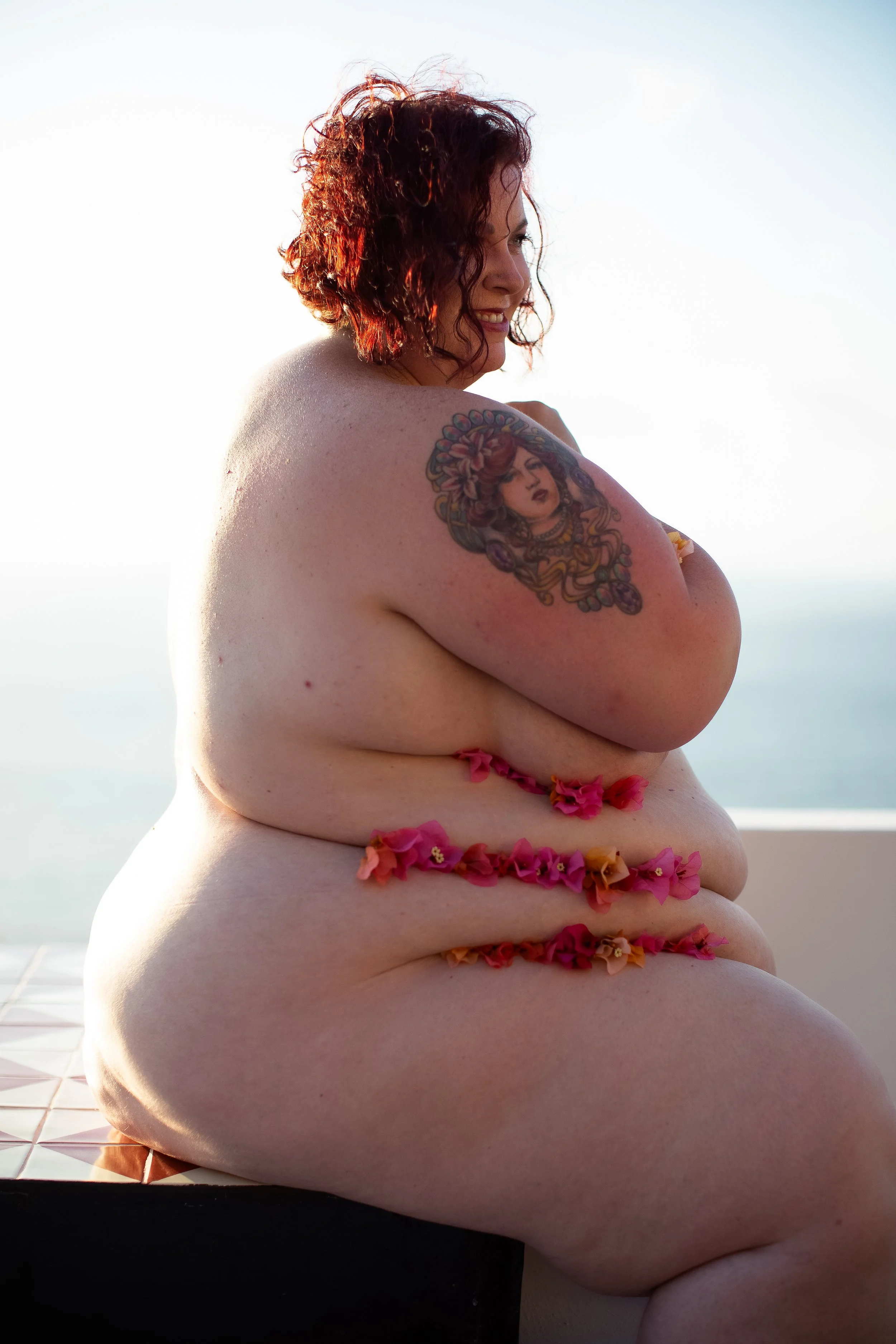 A woman with curly red hair and a tattoo on her arm, smiling, sitting topless with pink and orange flowers on her body, against a clear sky background.