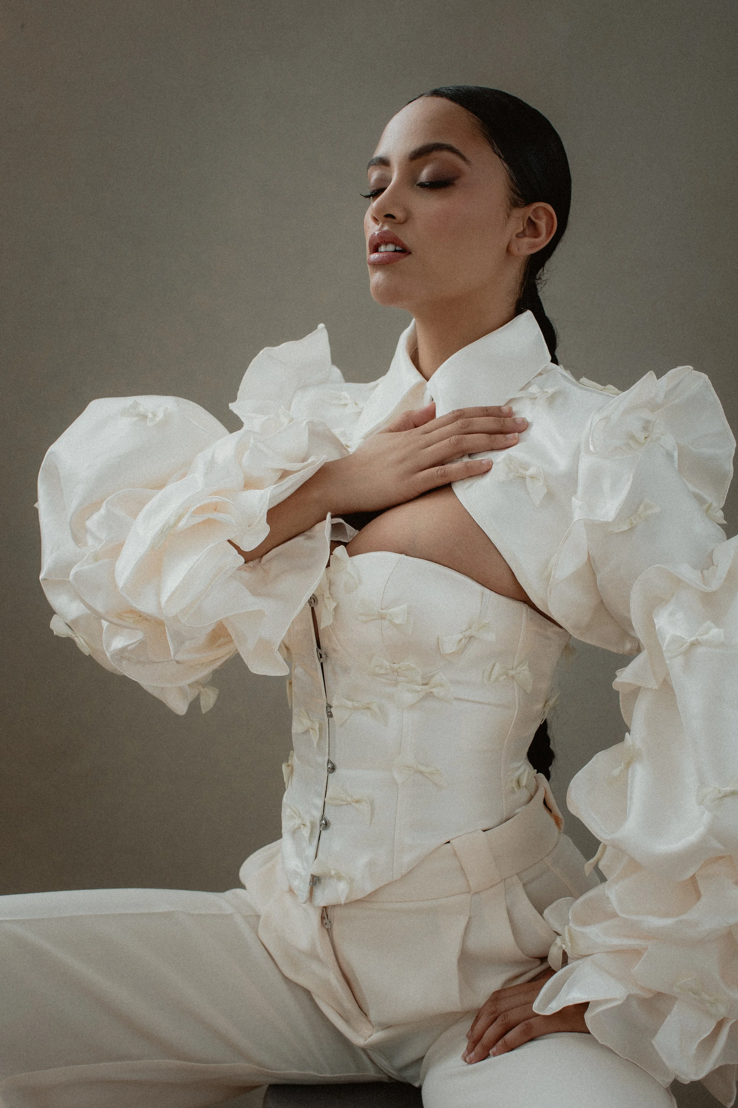 A woman with dark hair styled back, wearing a cream-colored outfit with voluminous, ruffled sleeves and a high collar, is posed with her eyes closed and hand on her chest against a neutral background.
