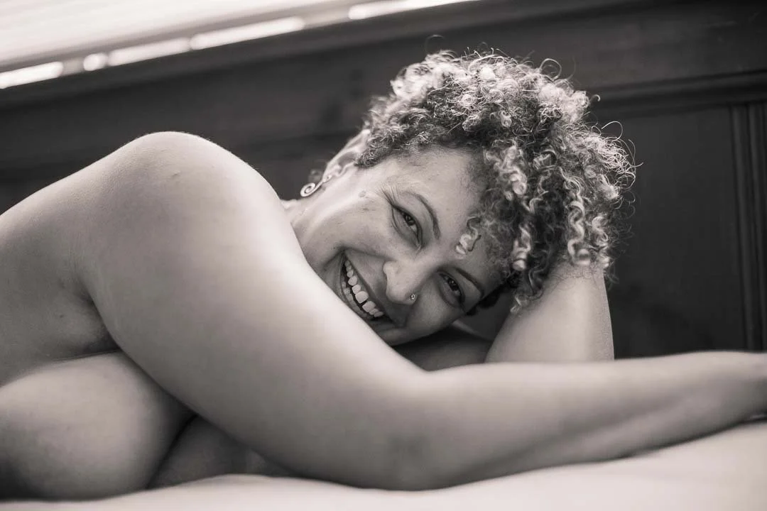 A woman with curly hair lying on a bed, smiling and looking at the camera.