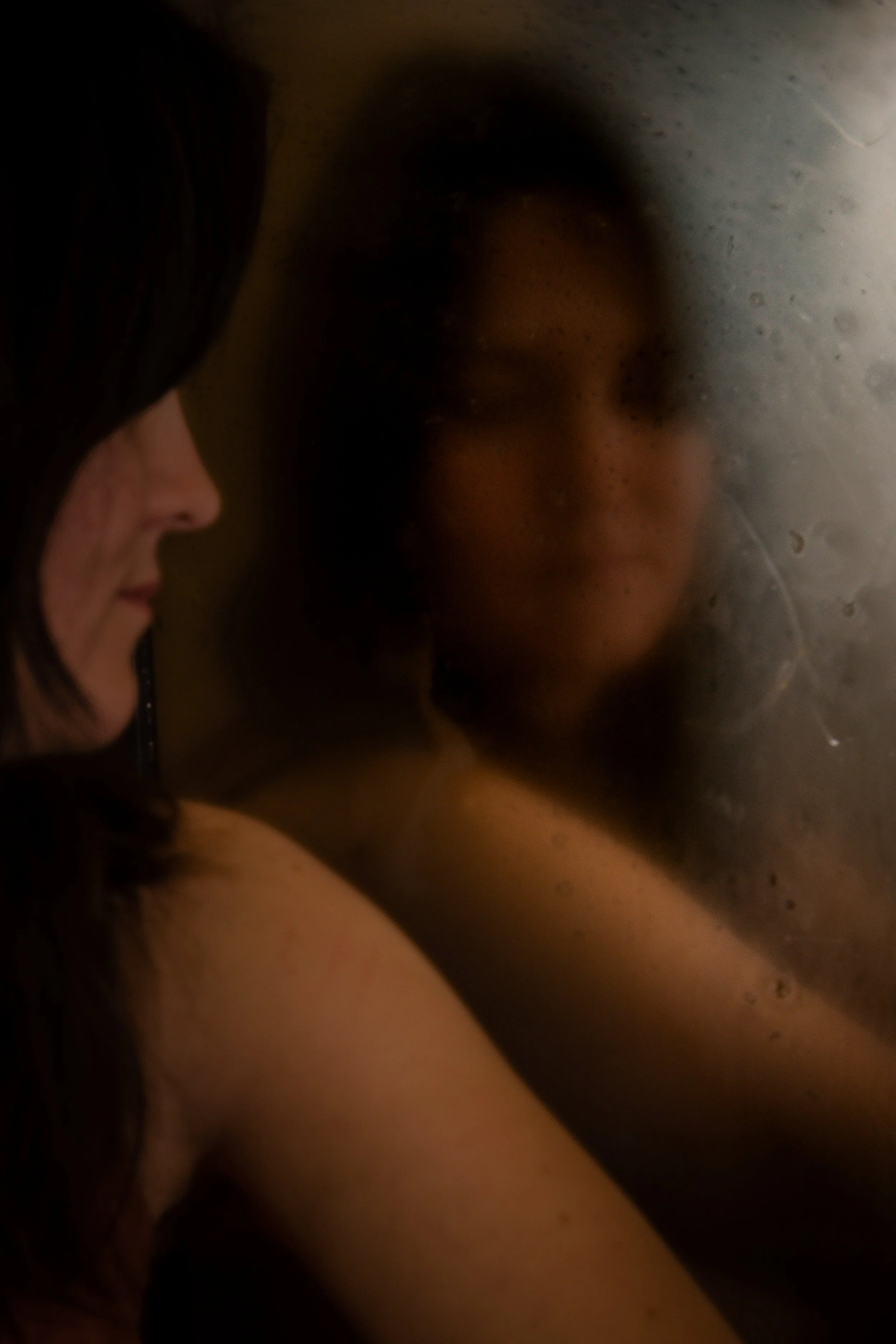 A close-up of two women's faces, one with dark hair and the other with lighter hair, with their foreheads and noses touching through a foggy or steamy surface.