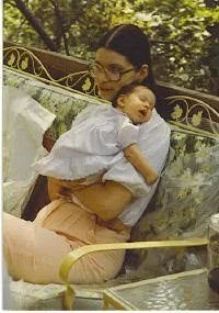 A woman holding a baby on her lap, sitting on a bench outdoors with trees in the background.