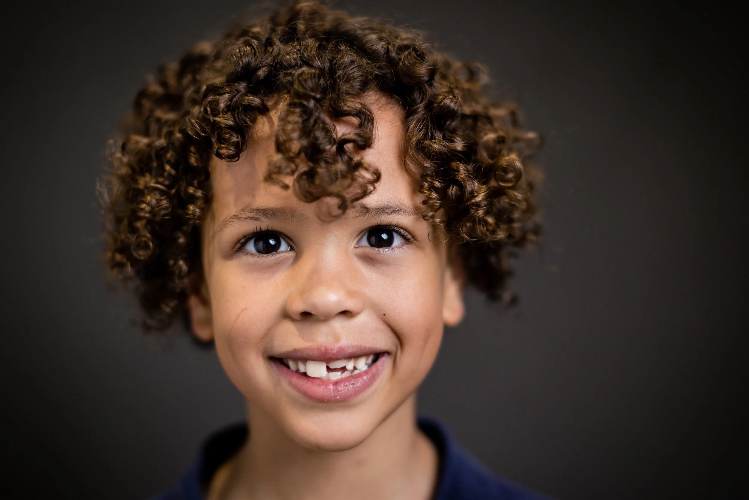 Close-up portrait of a smiling young child with curly hair and missing front teeth, against a dark background.