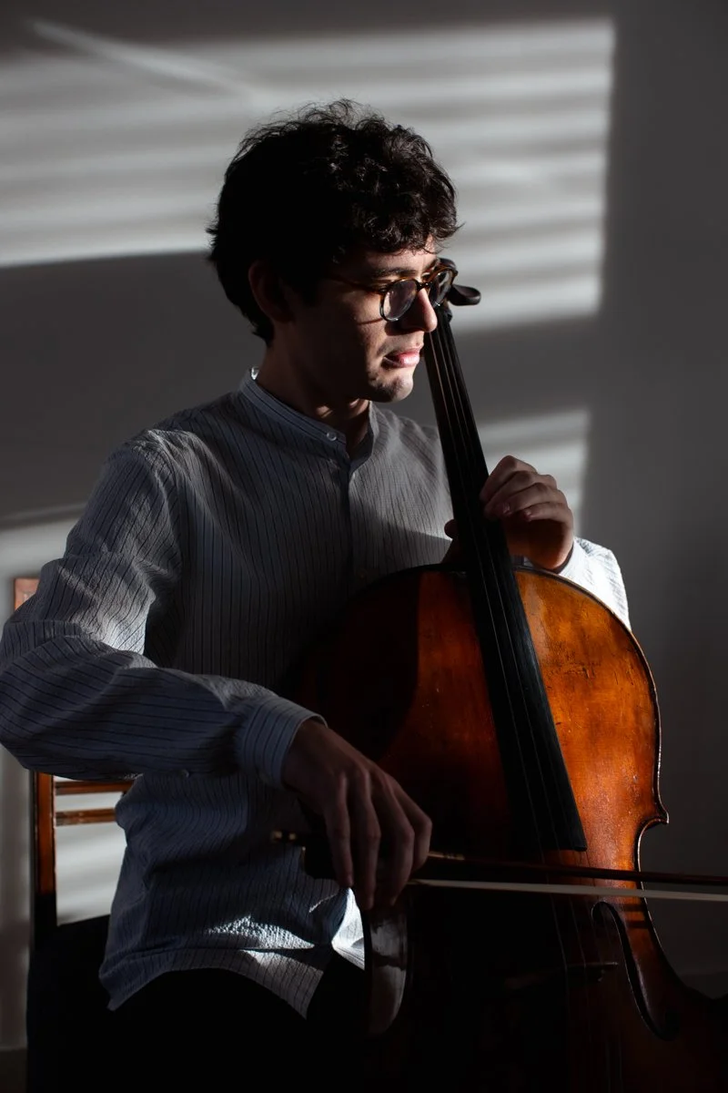 A man with dark curly hair, glasses, and a striped shirt is playing a wooden cello, with sunlight casting shadows through blinds on the wall behind him.