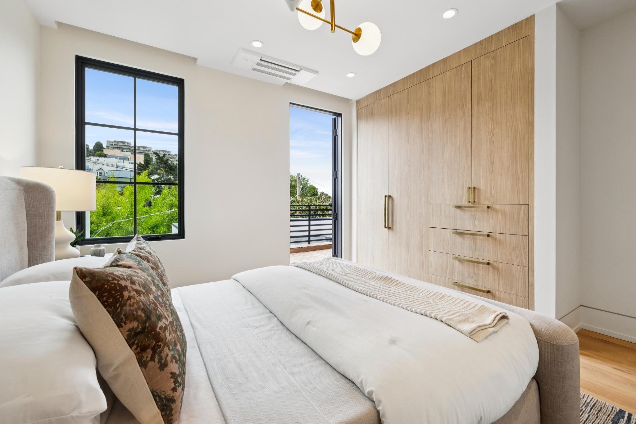 A modern bedroom with a white bed, decorative pillows, a bedside lamp, large windows showing green trees and a view of neighboring buildings, and a wooden closet with gold handles. There is a door leading to a balcony and a ceiling light fixture.
