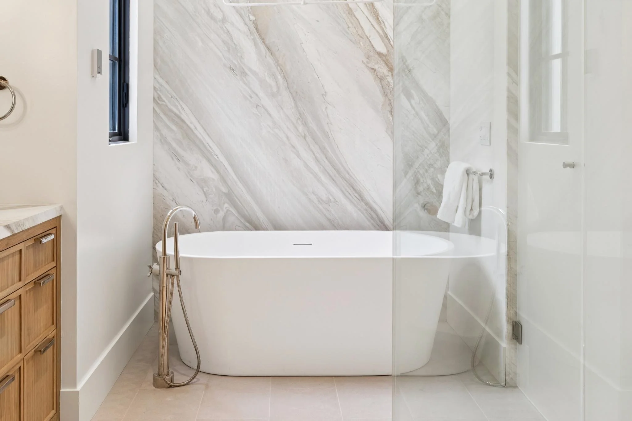 A modern bathroom with a walk-in shower, marble wall, freestanding bathtub, and a small window.