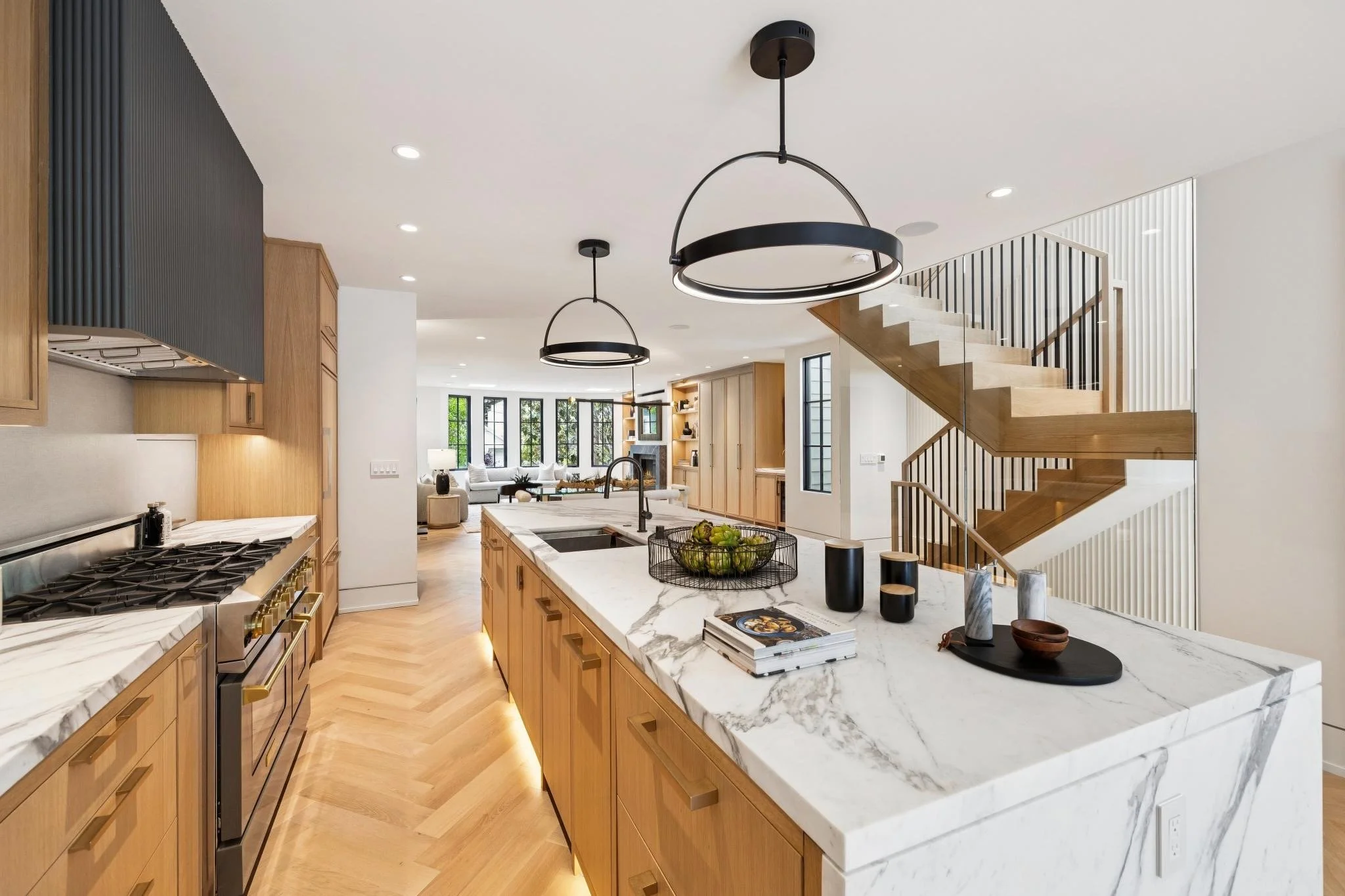 Modern open-concept kitchen with wooden cabinets, marble countertops, black and gold hardware, black pendant lights, and a staircase leading to the upper floor, with a living room visible in the background.