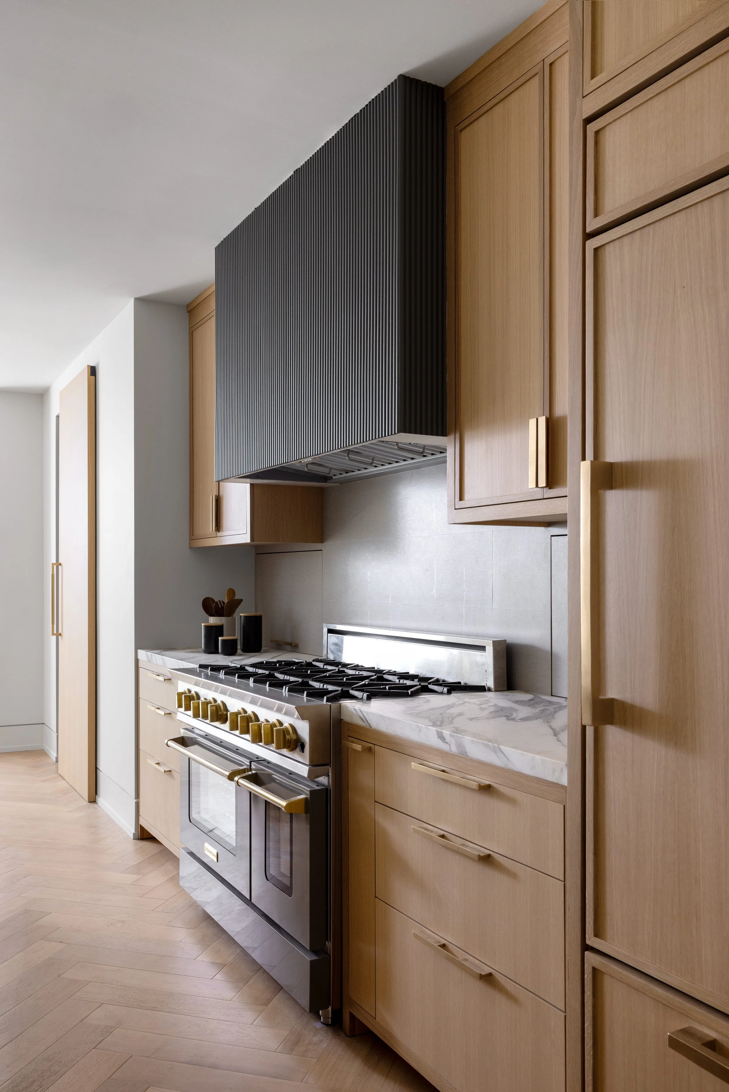 Modern kitchen with light wood cabinets, marble countertop, stainless steel oven, and black range hood.