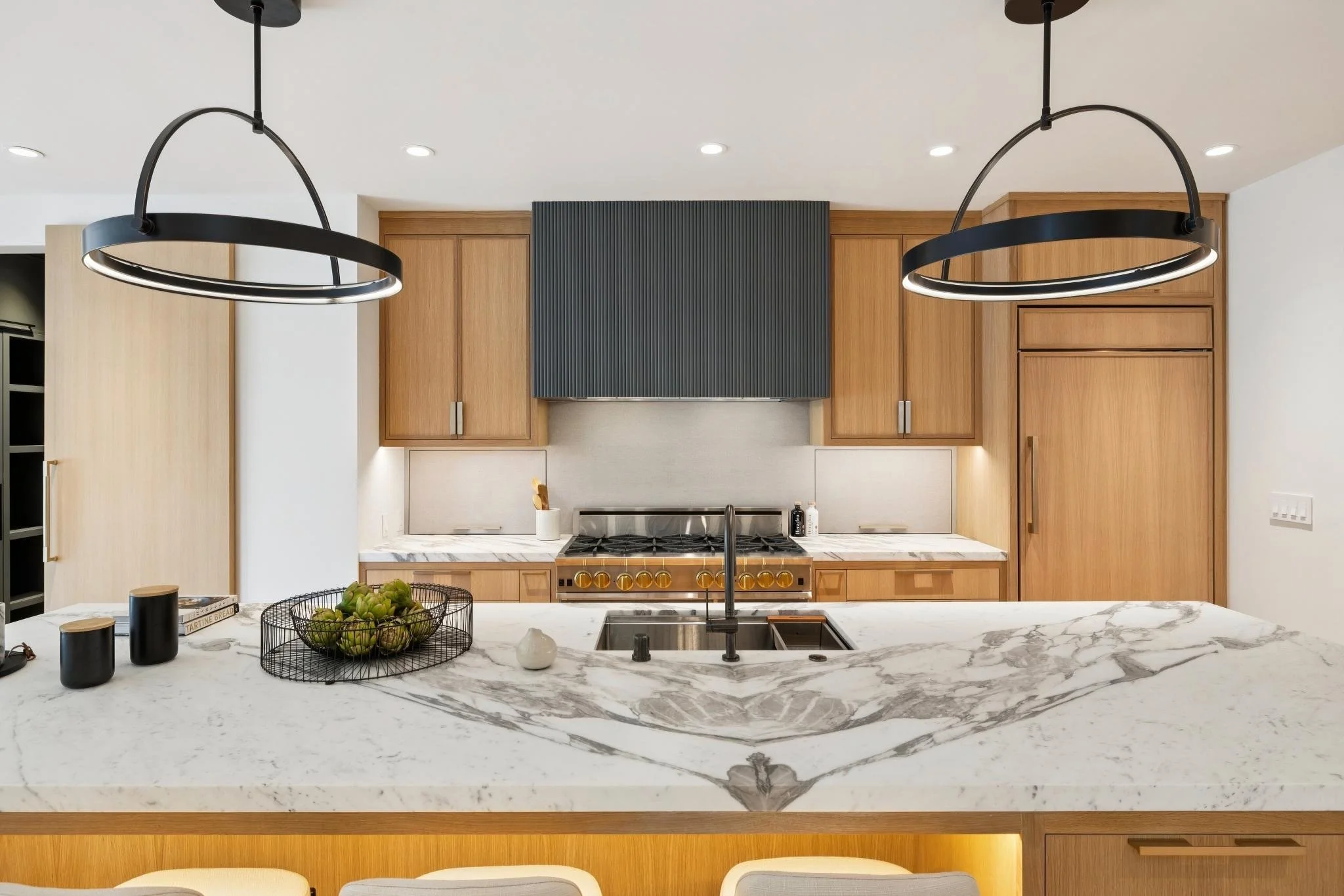Modern kitchen with wooden cabinets, marble island, black and gold stove, black pendant lights, and minimalist decor.