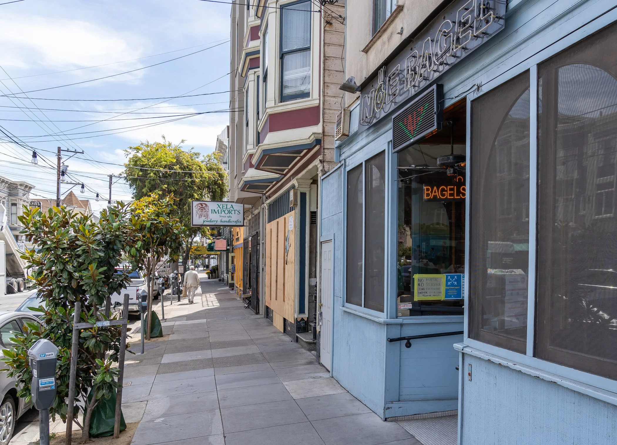 Street view with shops, including a blue storefront with neon signs for bagels and a green pepper symbol, and a sign for Xela Imports. Sidewalk with trees, parked cars, and pedestrians, under a partly cloudy sky.