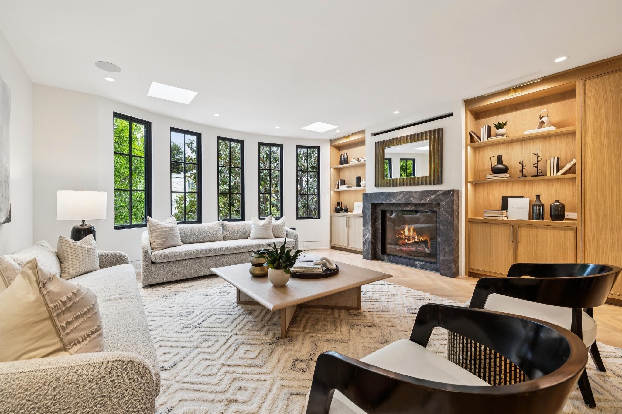 Living room with white walls and ceiling, multiple windows with trees outside, a coffee table with decorative items, beige sofas, a fireplace with a black marble surround, built-in wooden shelves with decorative objects, a mirror above the fireplace,