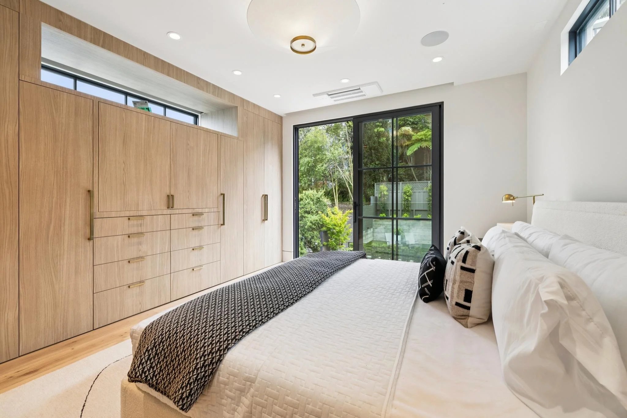 Modern bedroom with large wooden built-in closet, white bed with black and white pillows, black-framed sliding glass door leading to a green outdoor space, and a white ceiling with recessed lighting and a central ceiling fixture.