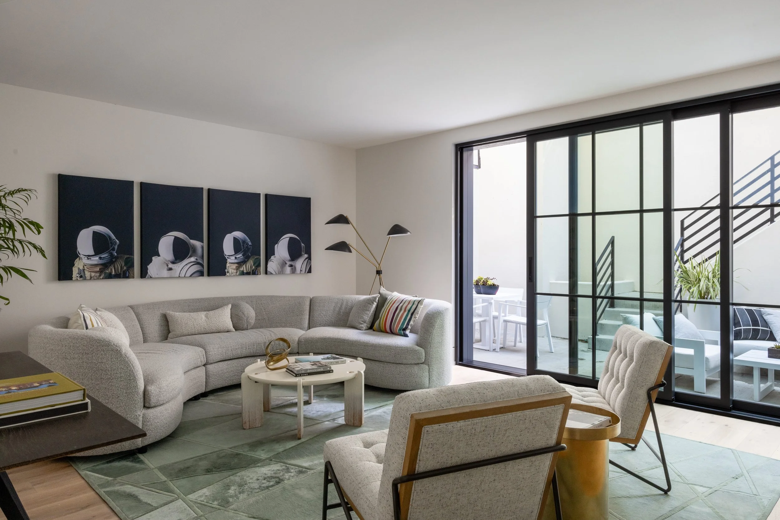 Modern living room with gray sectional sofa, abstract artwork of astronauts, glass sliding door to balcony with outdoor seating, and minimalistic decor.