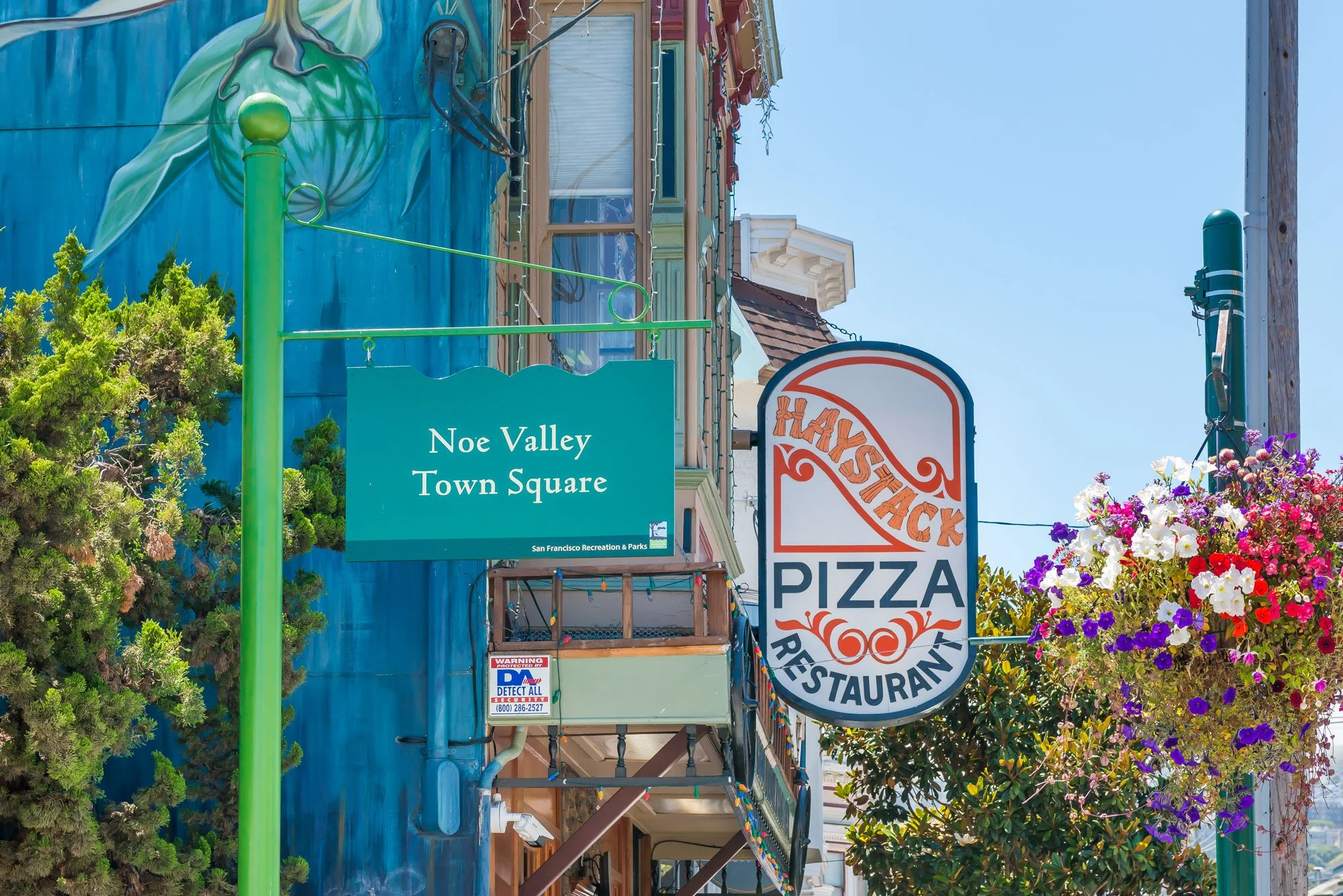 Street scene at Noe Valley Town Square with signs for Haystack Pizza Restaurant and colorful hanging flower baskets on a sunny day.