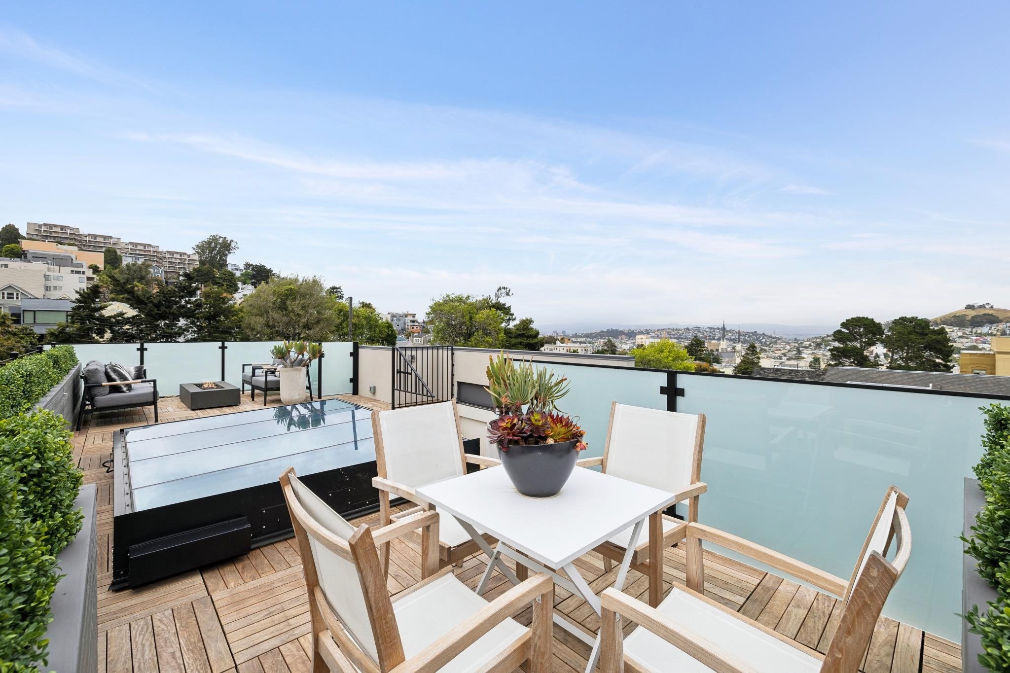 A backyard patio with outdoor furniture, including a white table, wooden chairs, and a black lounge, surrounded by potted plants, with a view of hills and cityscape in the distance under a partly cloudy sky.