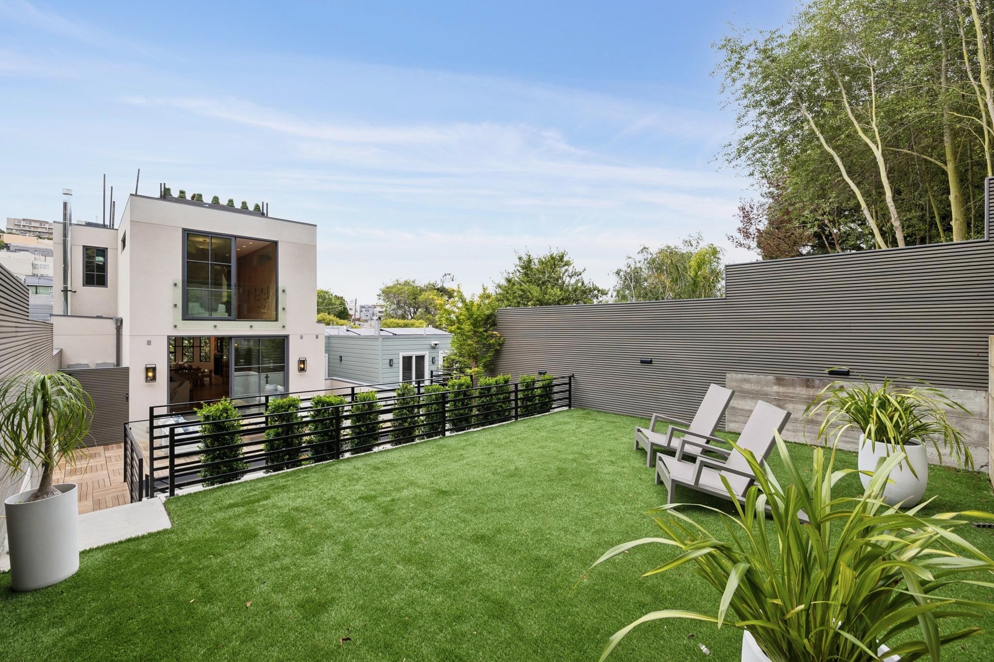 Modern backyard with a green lawn, potted plants, lounge chairs, and a sleek fence, overlooking neighboring houses and trees under a clear blue sky.