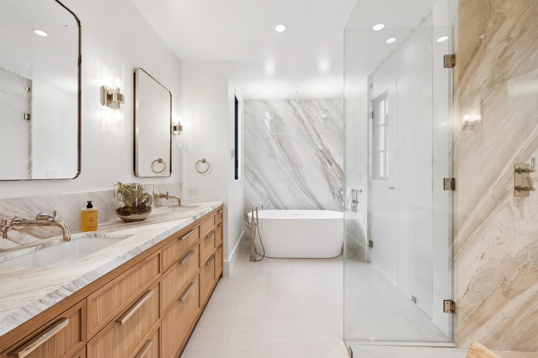 Modern bathroom with wooden vanity, marble countertop, large mirrors, a bathtub, and a glass-enclosed shower with marble walls.