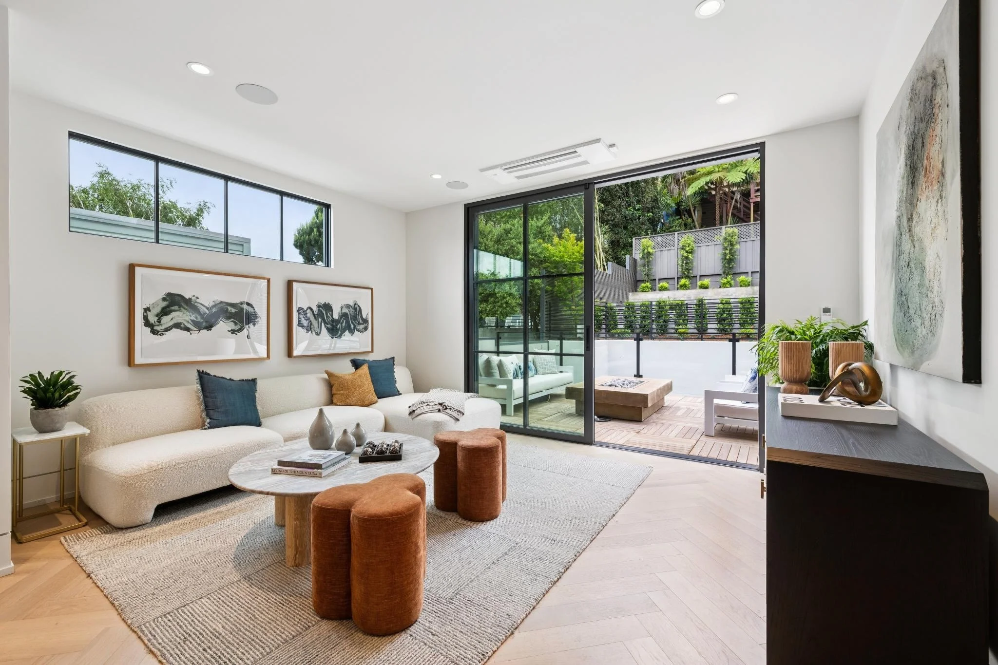 Modern living room with white walls, a beige sofa with blue and mustard pillows, a round coffee table, orange stools, indoor plants, abstract black and white artwork, and large sliding glass doors opening to an outdoor patio with greenery.
