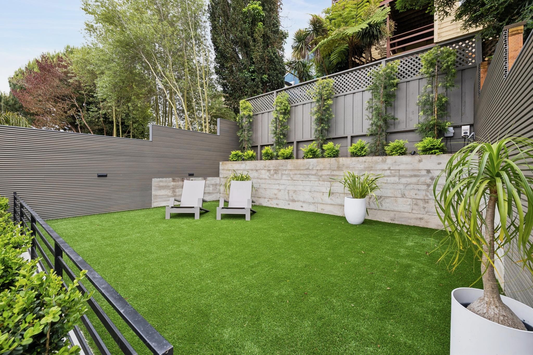 A modern backyard with a well-maintained artificial grass lawn, two white lounge chairs, potted plants, a wooden privacy fence, and tall trees and shrubs surrounding the area.