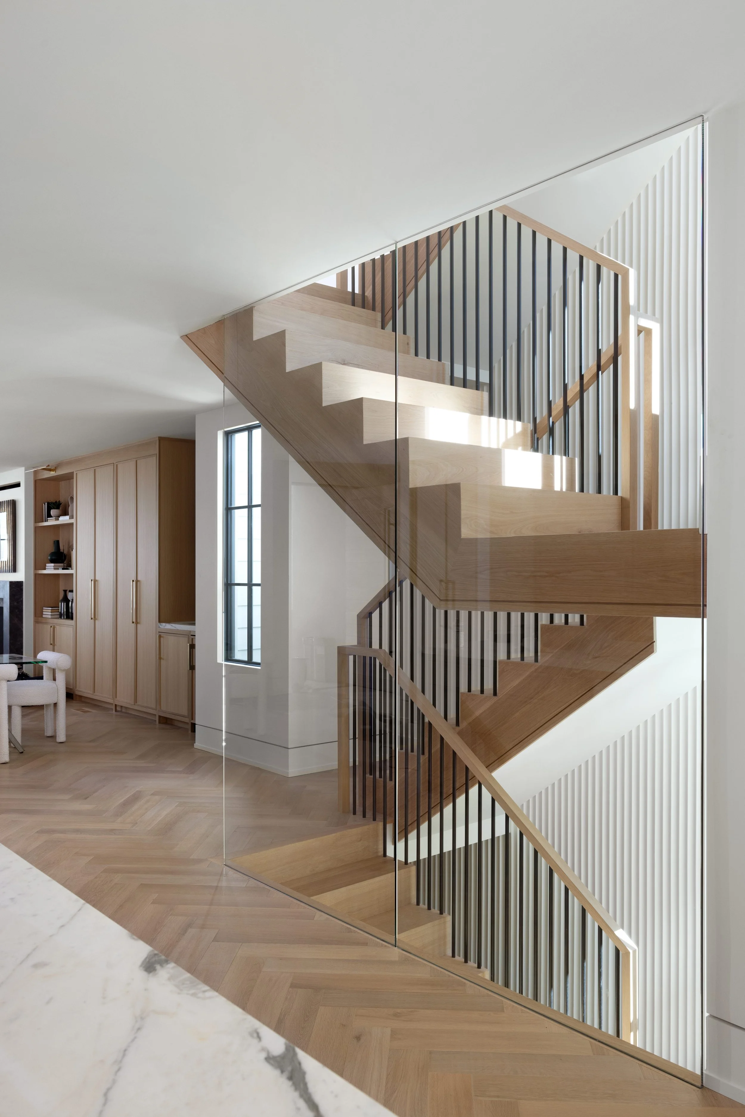 Interior view of a modern wooden staircase with vertical black metal railings, reflected in a large mirror on a white wall, in a bright, minimalistic home.