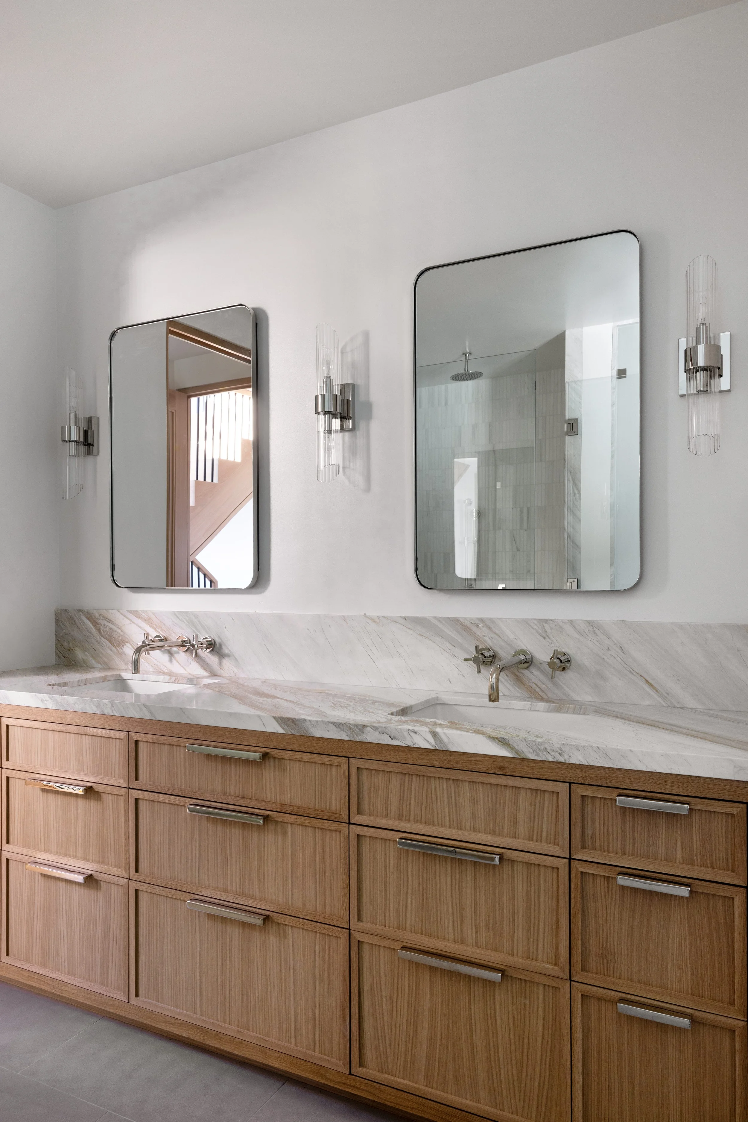 Modern bathroom with a double vanity, marble countertop, and two rectangular mirrors, illuminated by wall-mounted sconces, with a view of a shower in the background.