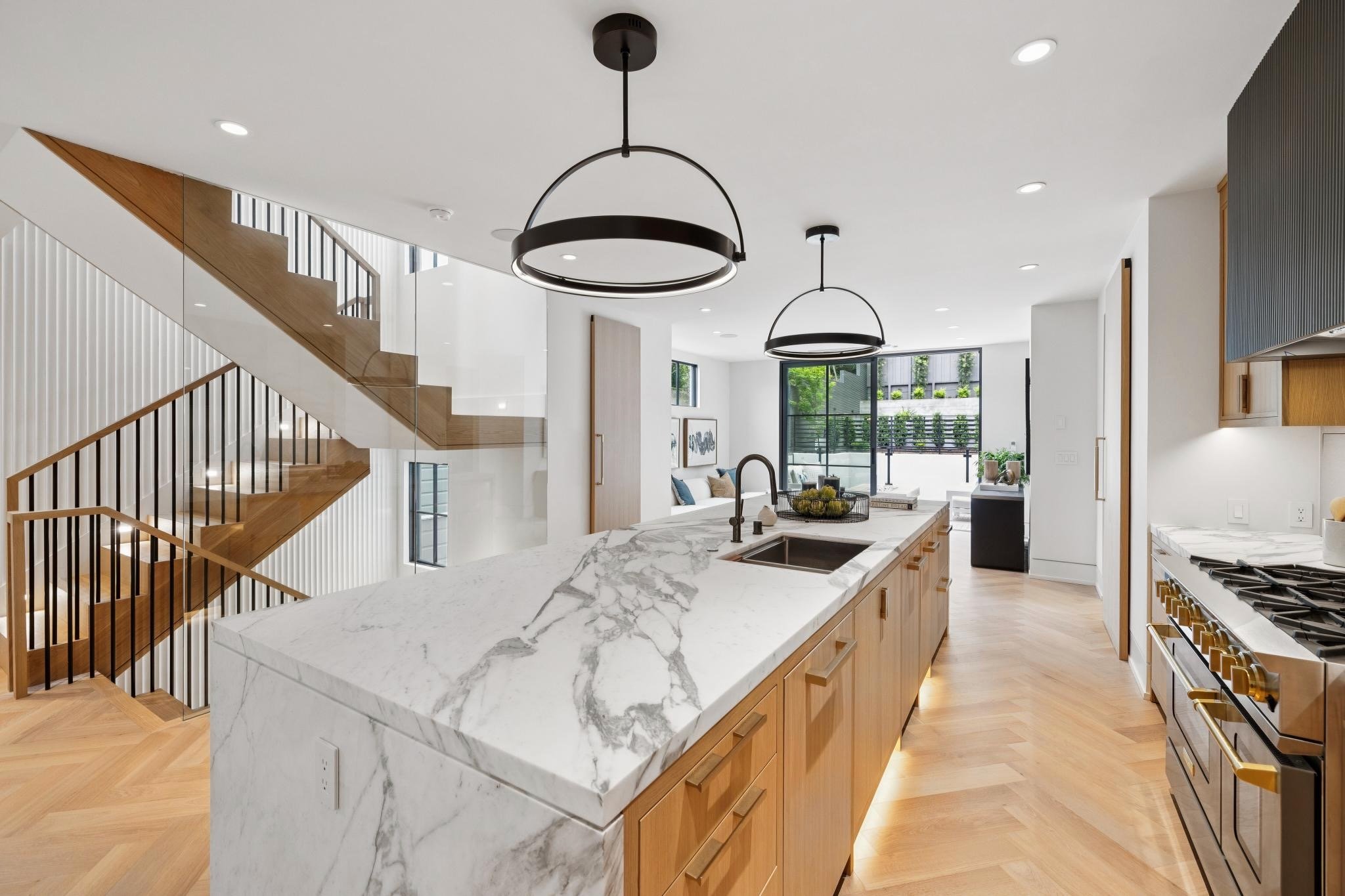 Modern kitchen with white marble island, wooden cabinets, black circular pendant lights, stainless steel oven, and large windows with outdoor greenery.