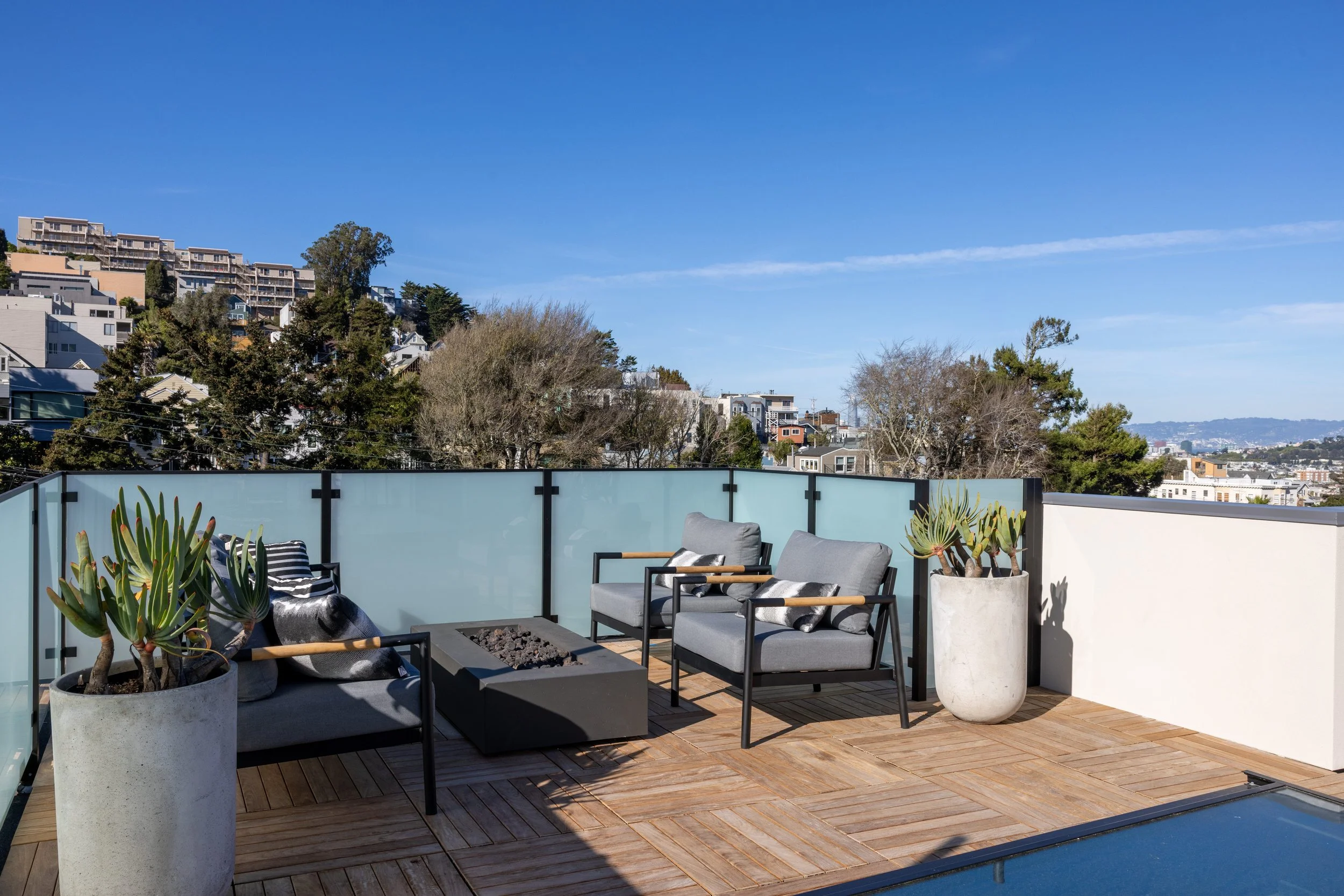 Outdoor rooftop terrace with two gray armchairs, a black fire pit, large potted succulents, and glass safety barriers, overlooking a cityscape under a clear blue sky.