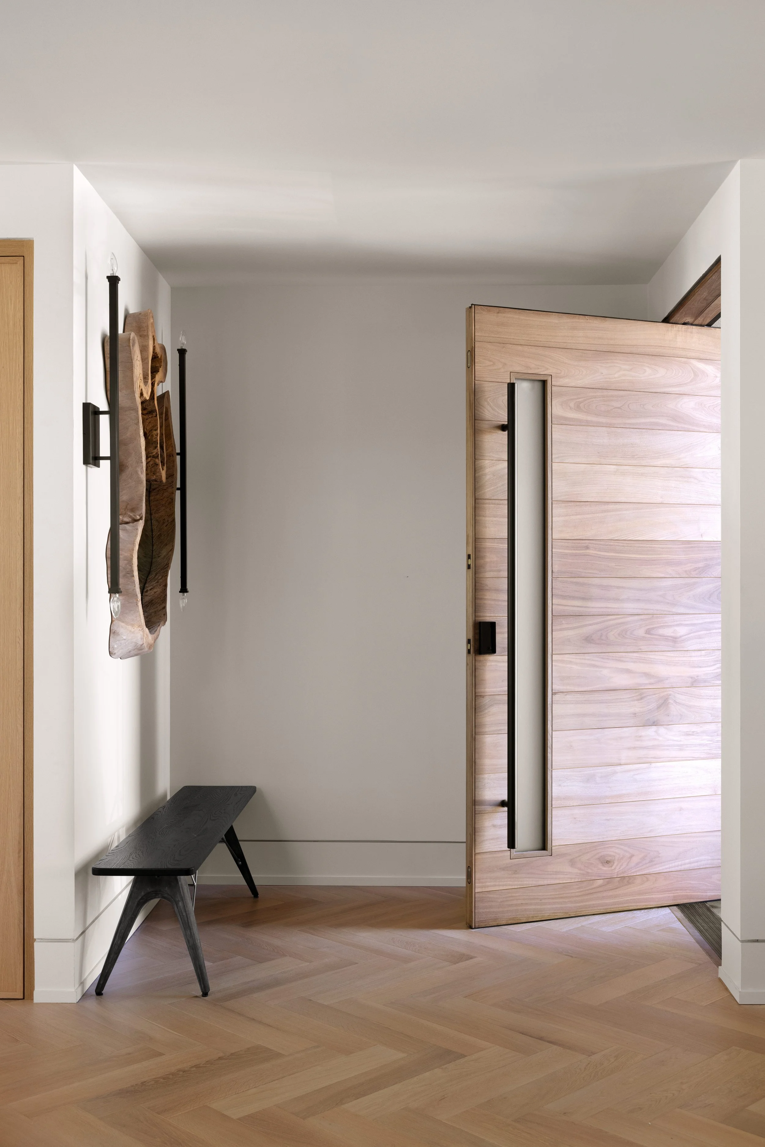 A modern interior with a wooden door, light-colored walls, a small black bench, and decorative wall art.