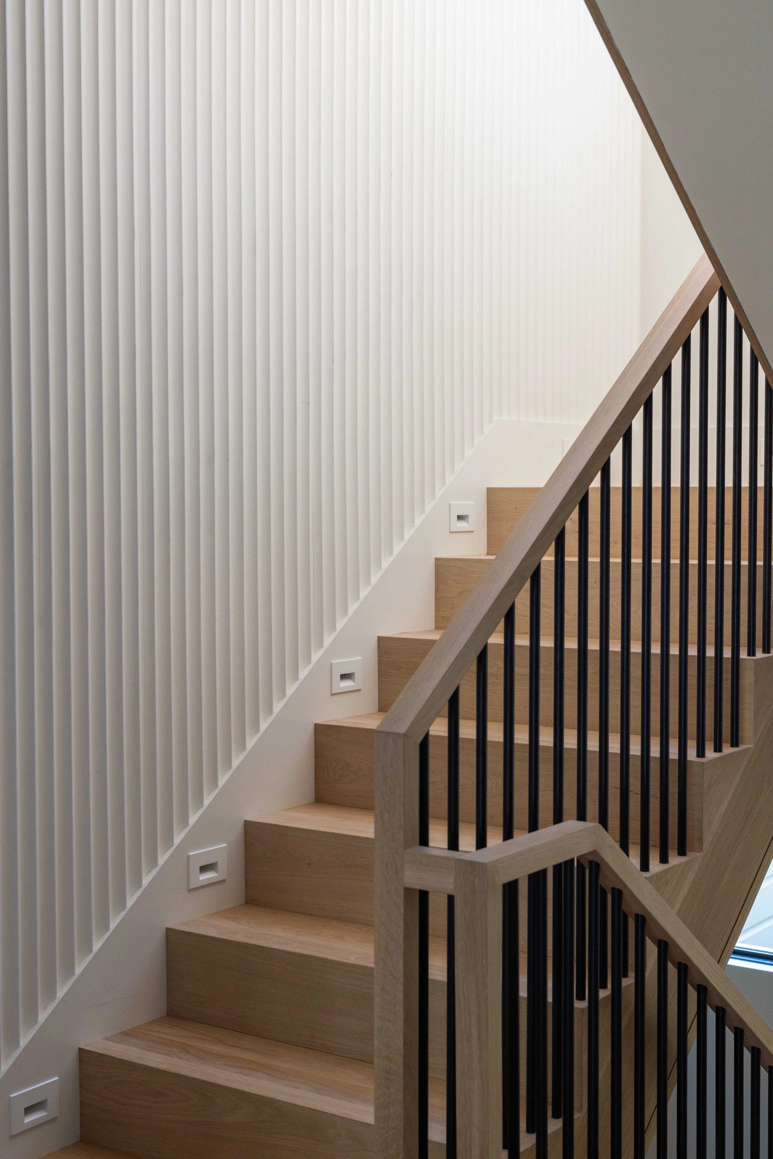 Modern staircase with wooden steps, black metal railing, and a textured white wall with vertical lines.