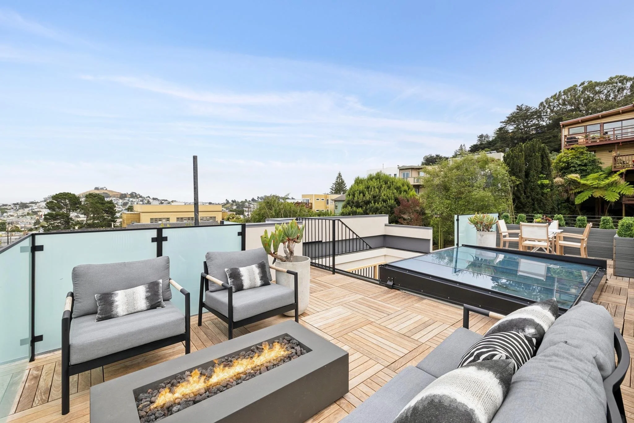 A modern outdoor balcony with comfortable seating, a fire feature, and a scenic view of trees and rooftops under a blue sky.