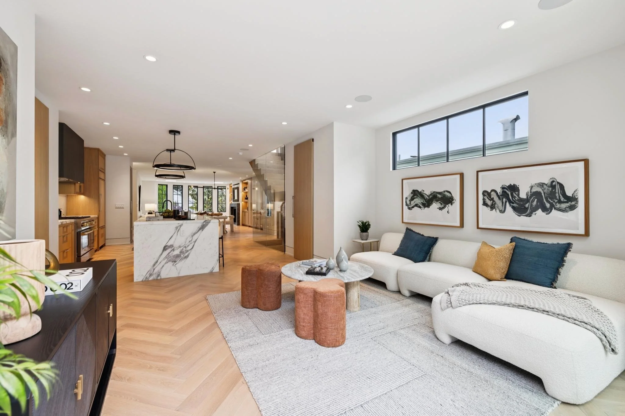 A modern, open-concept living room with a white sofa, colorful cushions, abstract black and white art on the wall, a marble coffee table, and a large window providing natural light.