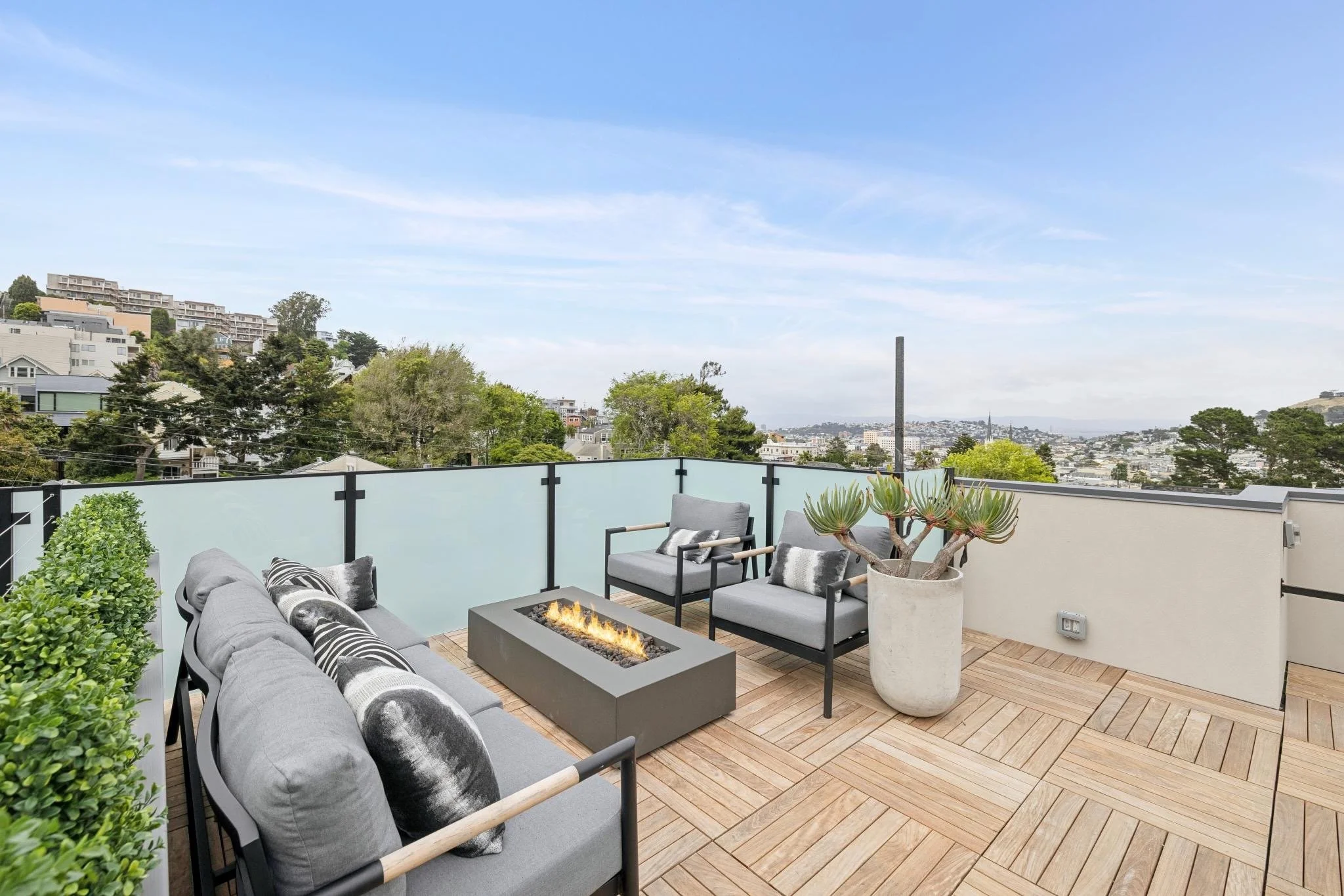 View of a spacious outdoor balcony with a seating area, fire pit, and potted succulent, overlooking a hillside neighborhood and blue sky.